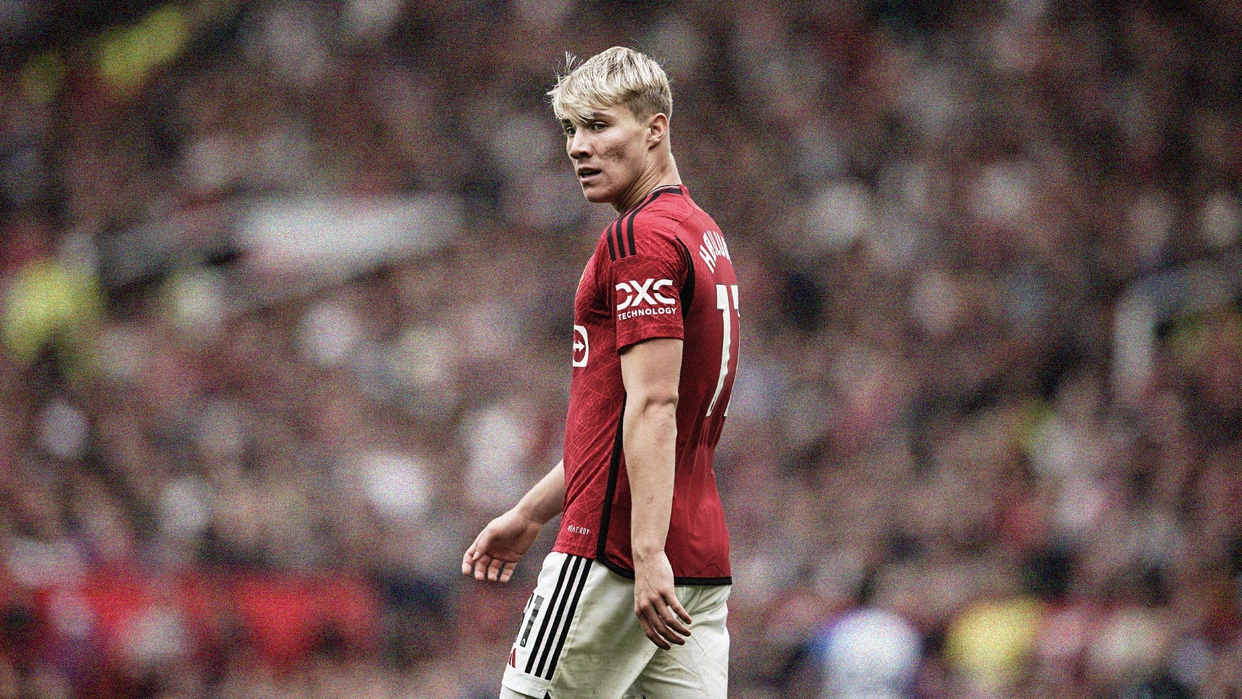 A photo of Rasmus Højlund playin for Manchester United. He's the only person in shot, looking over his left shoulder, with a blurred crowd in the background.