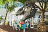 A person sits at a table underneath a statue of a dinosaur named Stan with a Google logo hanging from his teeth outside a glass building at Google HQ