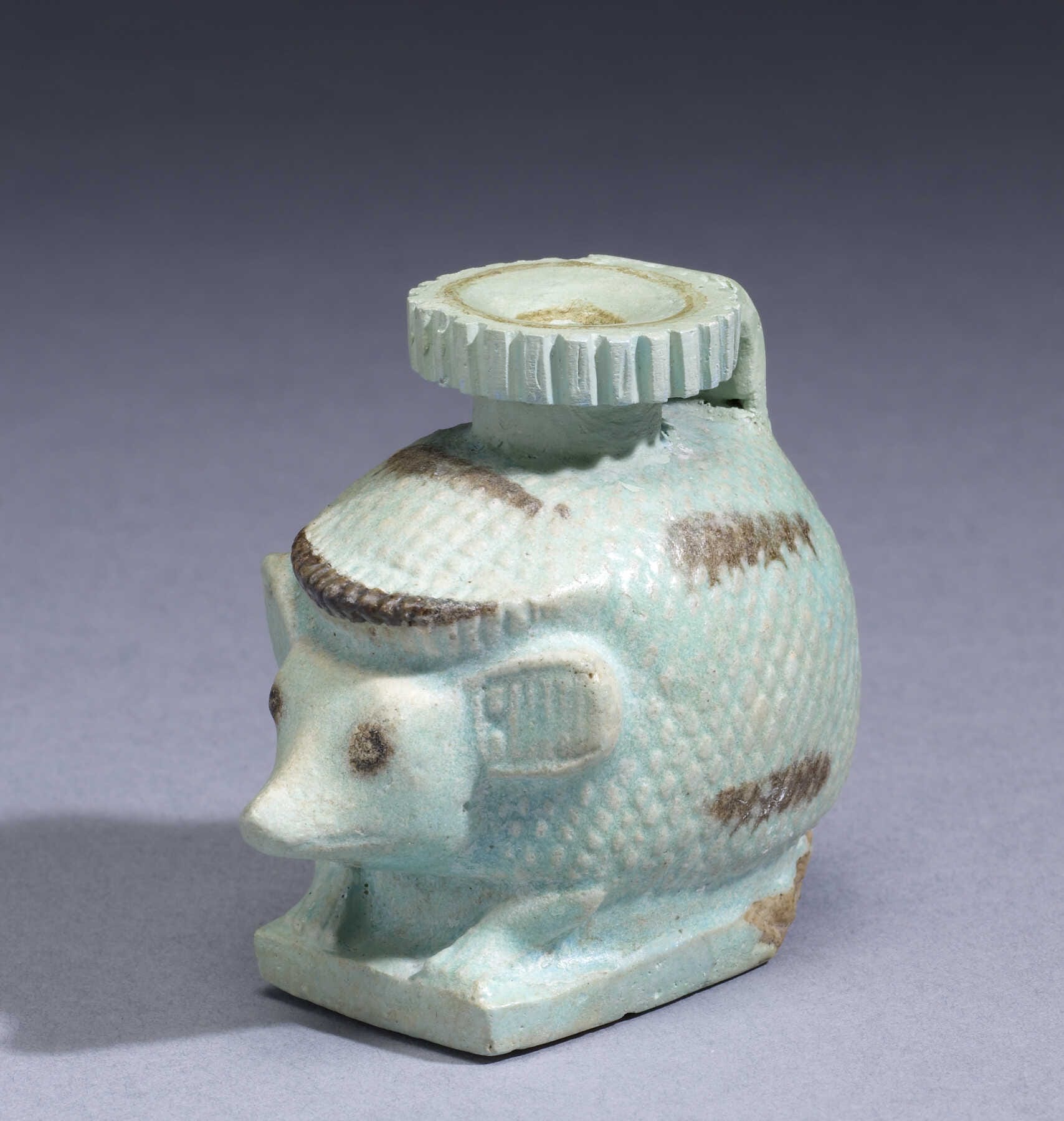 A small Greco-Egyptian perfume bottle (aryballos) shaped like a hedgehog, standing on a rectangular base. Its prickly body is cross-hatched, with details in dark glaze showing eyes, snout, and upright ears