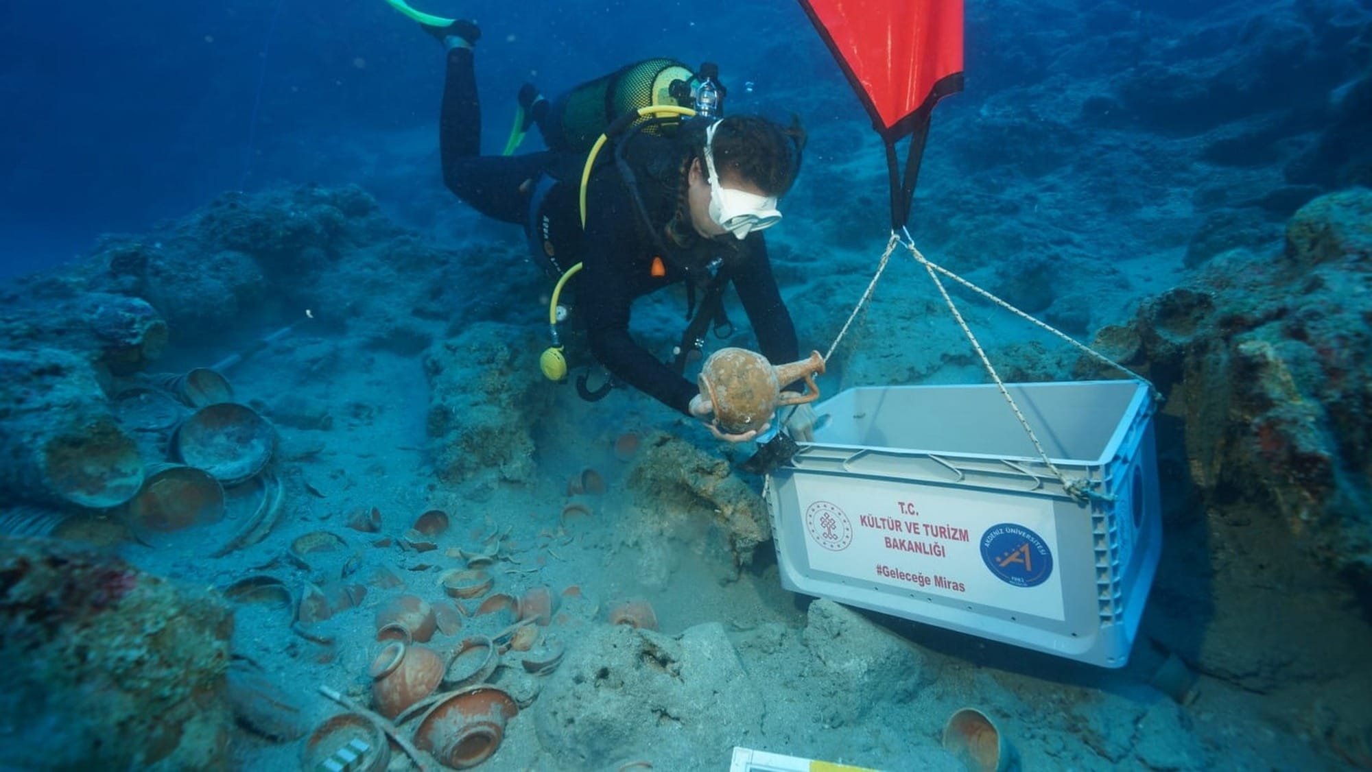 A diver holding a ceramic vessel beside a suspended recovery crate, with ancient dishes on the Adrasan seabed.
