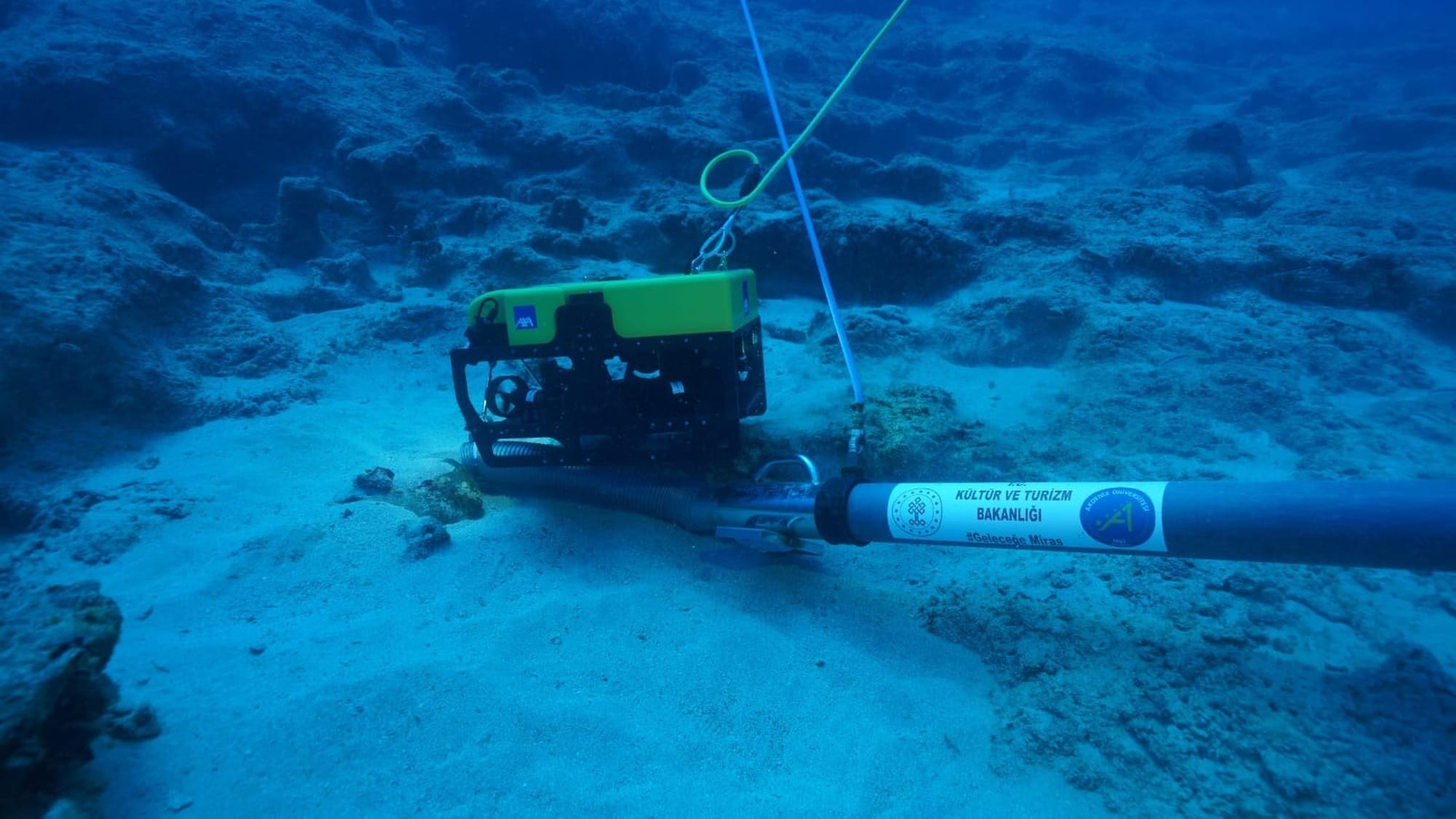 A tethered underwater robot and excavation equipment beside rocks and sand at the Adrasan wreck site.