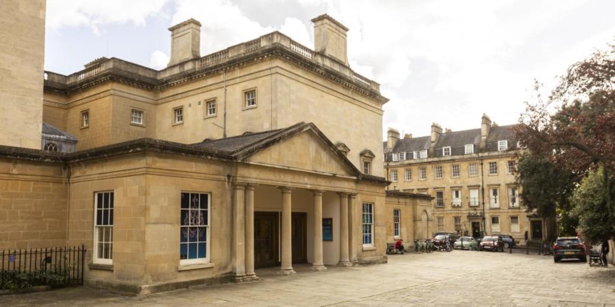Stone frontage and entrance portico of Bath Assembly Rooms beside a paved square.