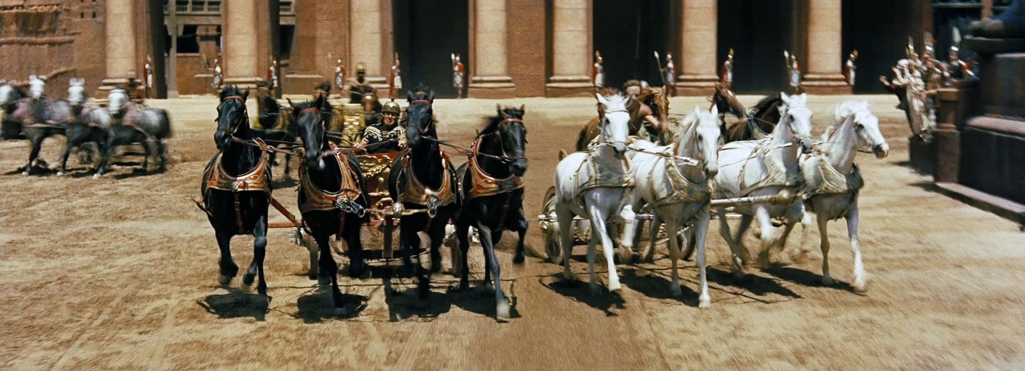 Black and white teams of horses race side by side in the arena in a colour frame from Ben-Hur (1959).
