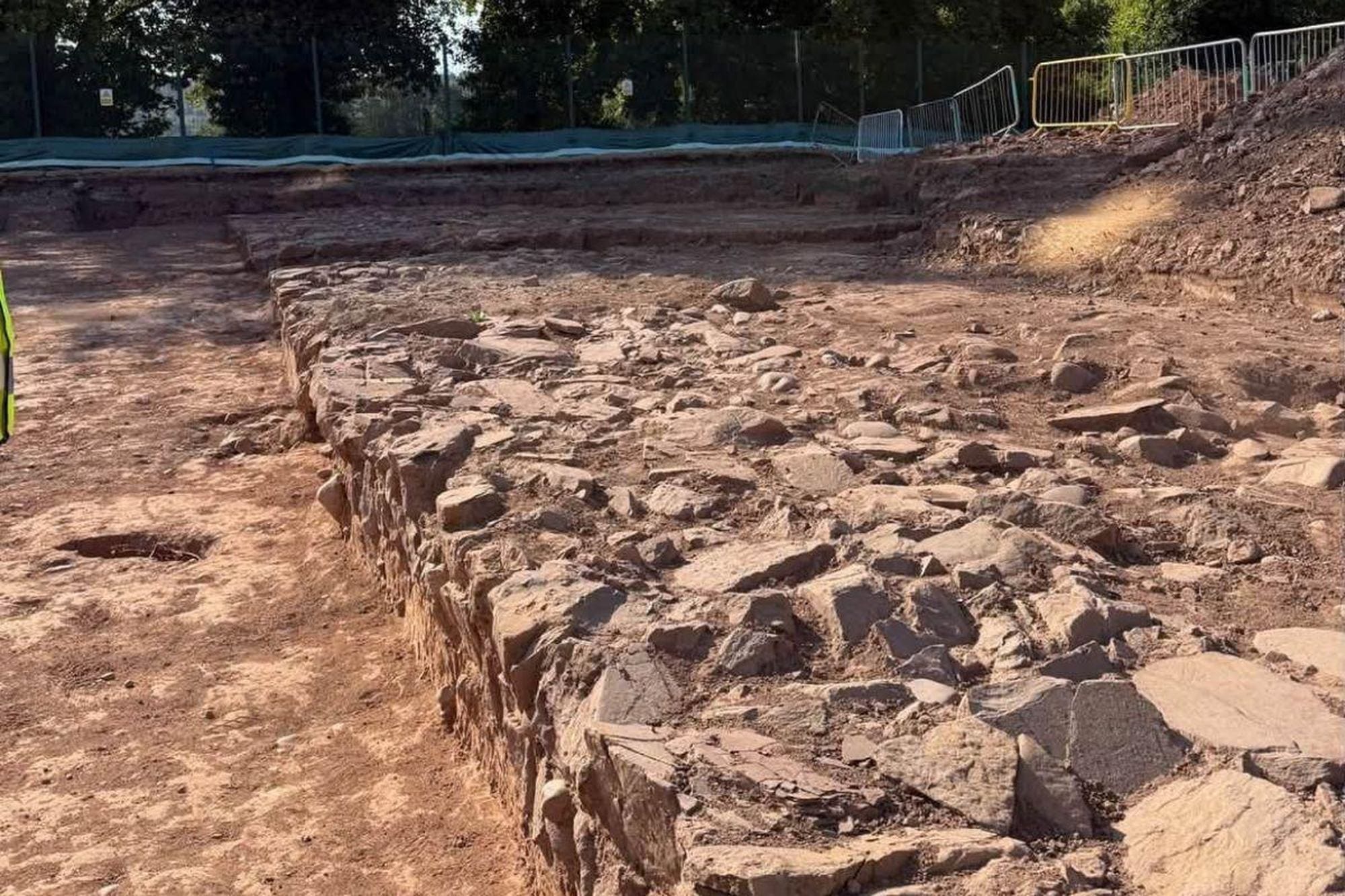 Wide excavation exposing a low stone structure and surrounding soil at Caerleon Comprehensive School.