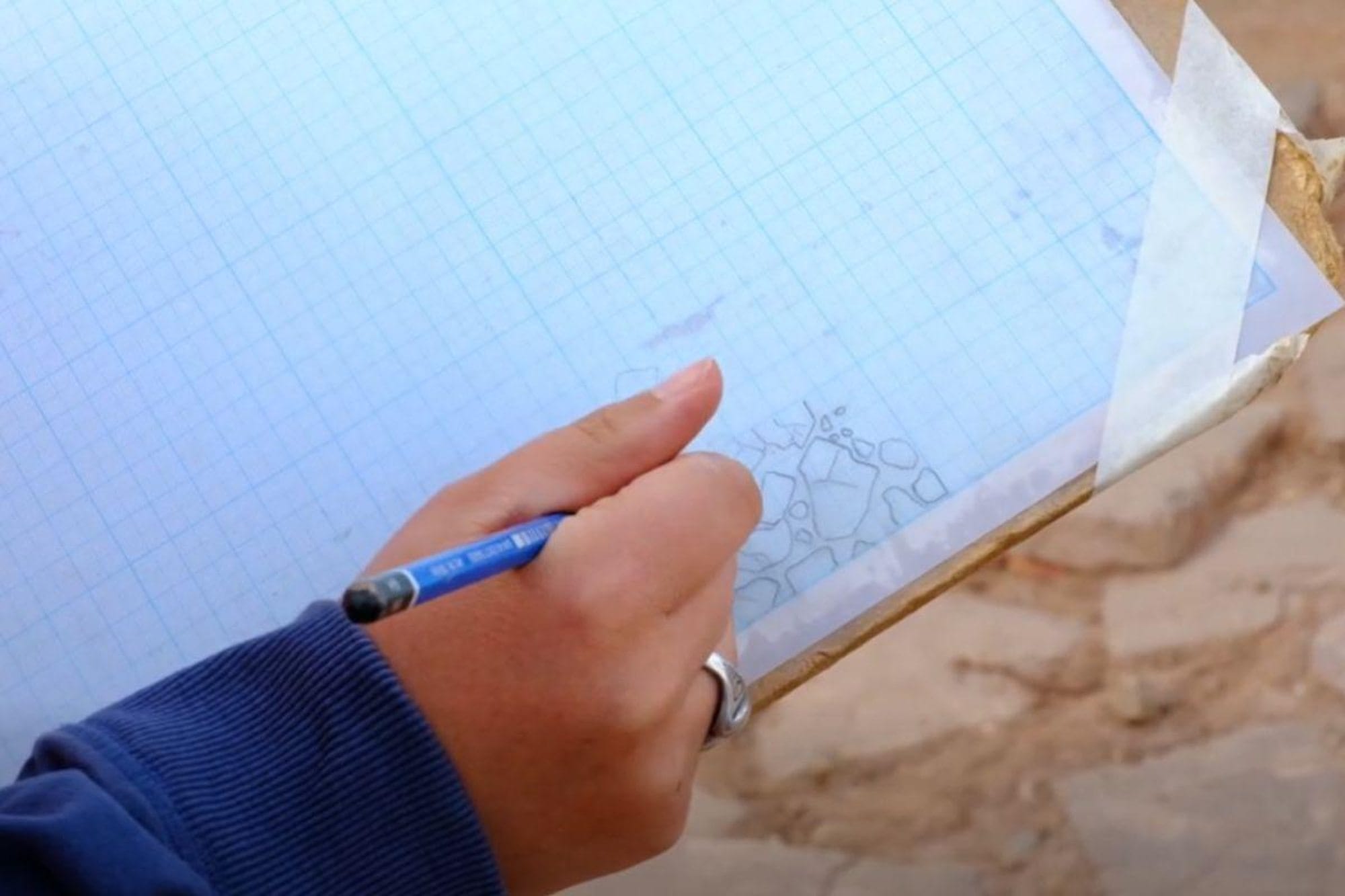 A hand uses a pencil to draw excavated stones on blue graph paper at the Caerleon school site.