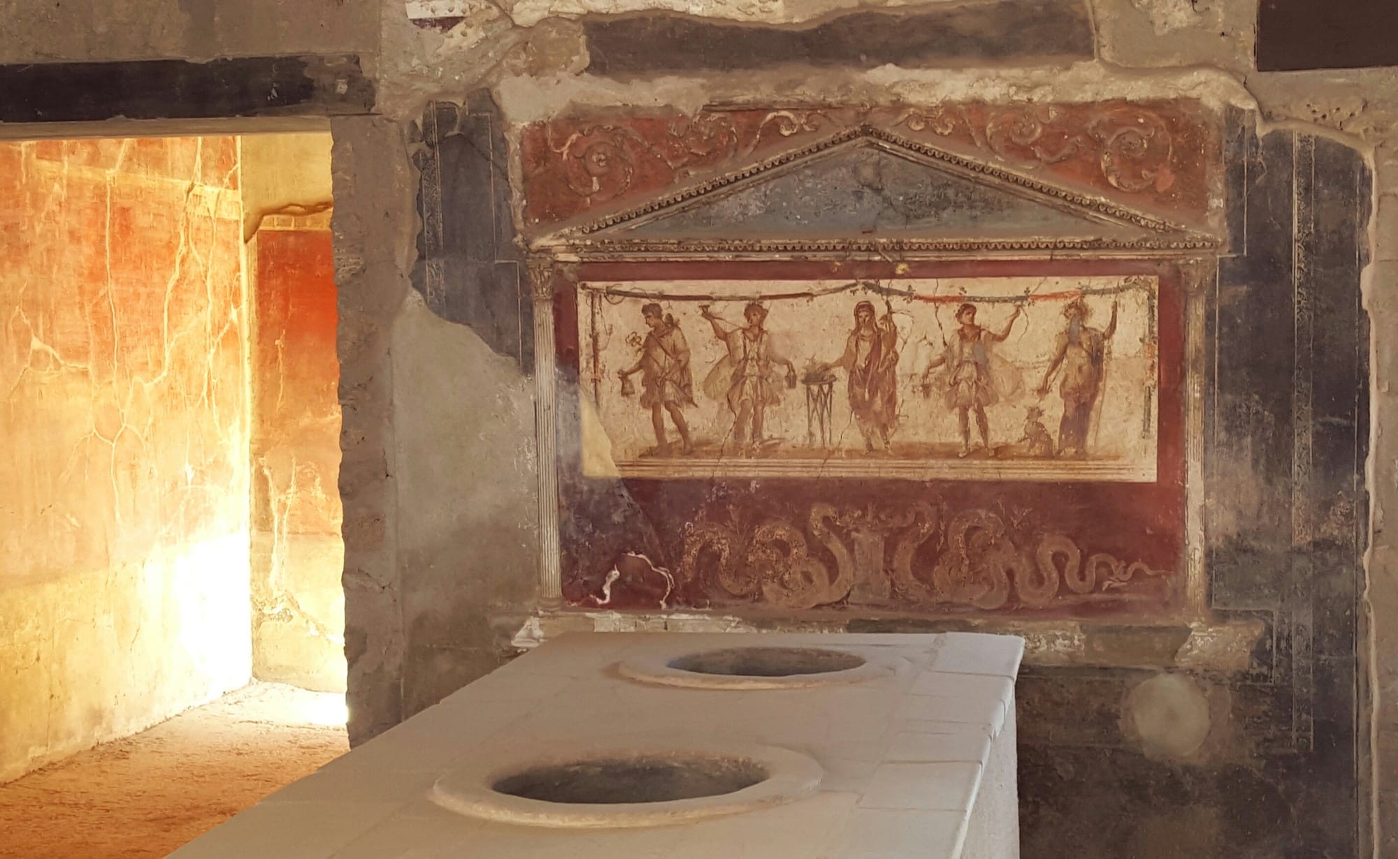 A marble-clad Roman food counter with round vessel openings stands before a painted shrine in Pompeii.