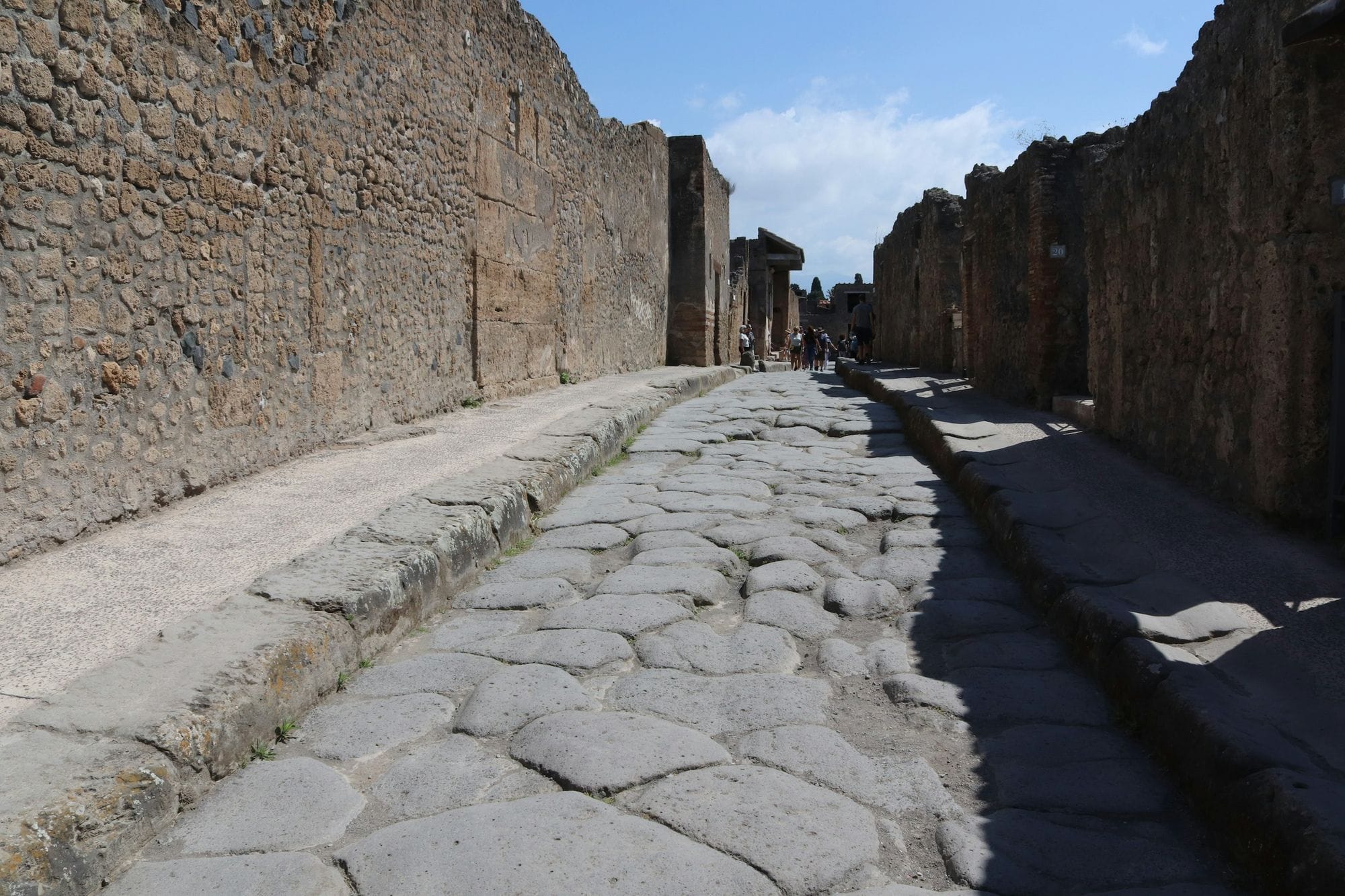 A stone-paved street runs between ancient building walls under a blue sky at Pompeii.