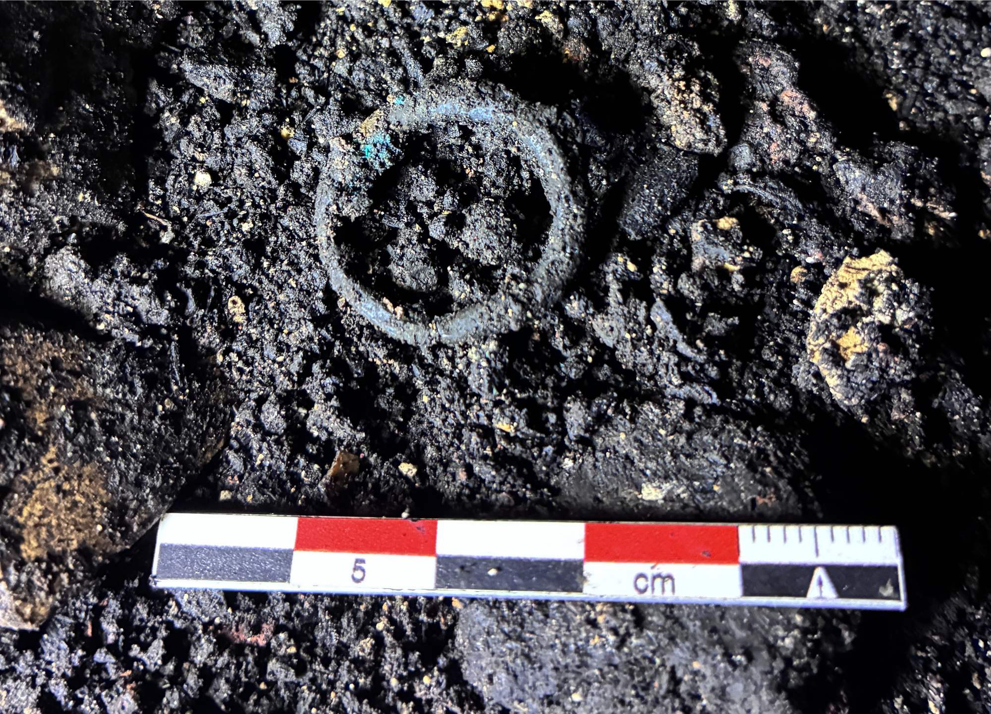 A second bronze ring lies on the cave surface beside a red, white and black archaeological scale.
