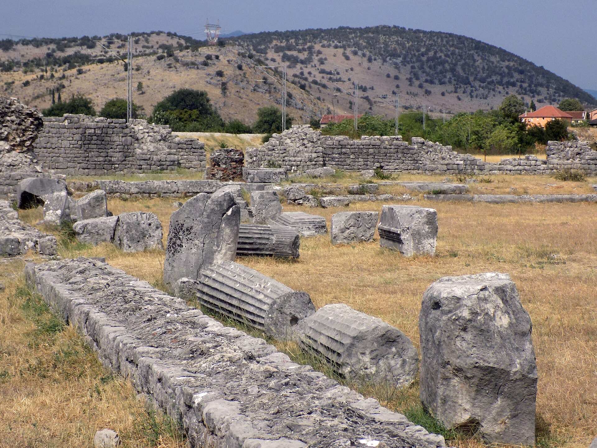 Stone walls and architectural blocks scattered across the ruins of Roman Doclea near Podgorica, with low hills behind.