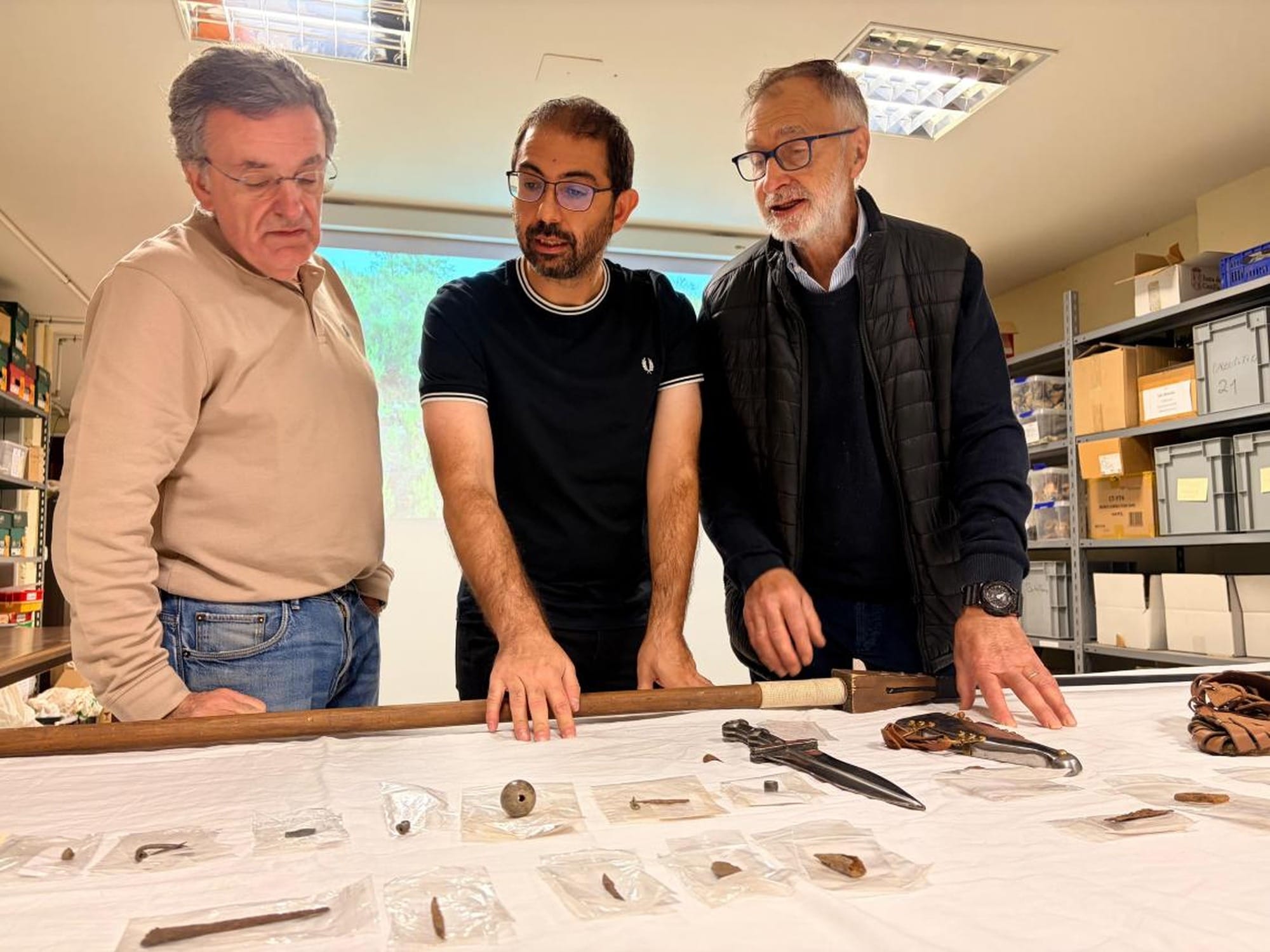 Three men examining small archaeological finds laid out on a table, including metal objects in labelled bags.