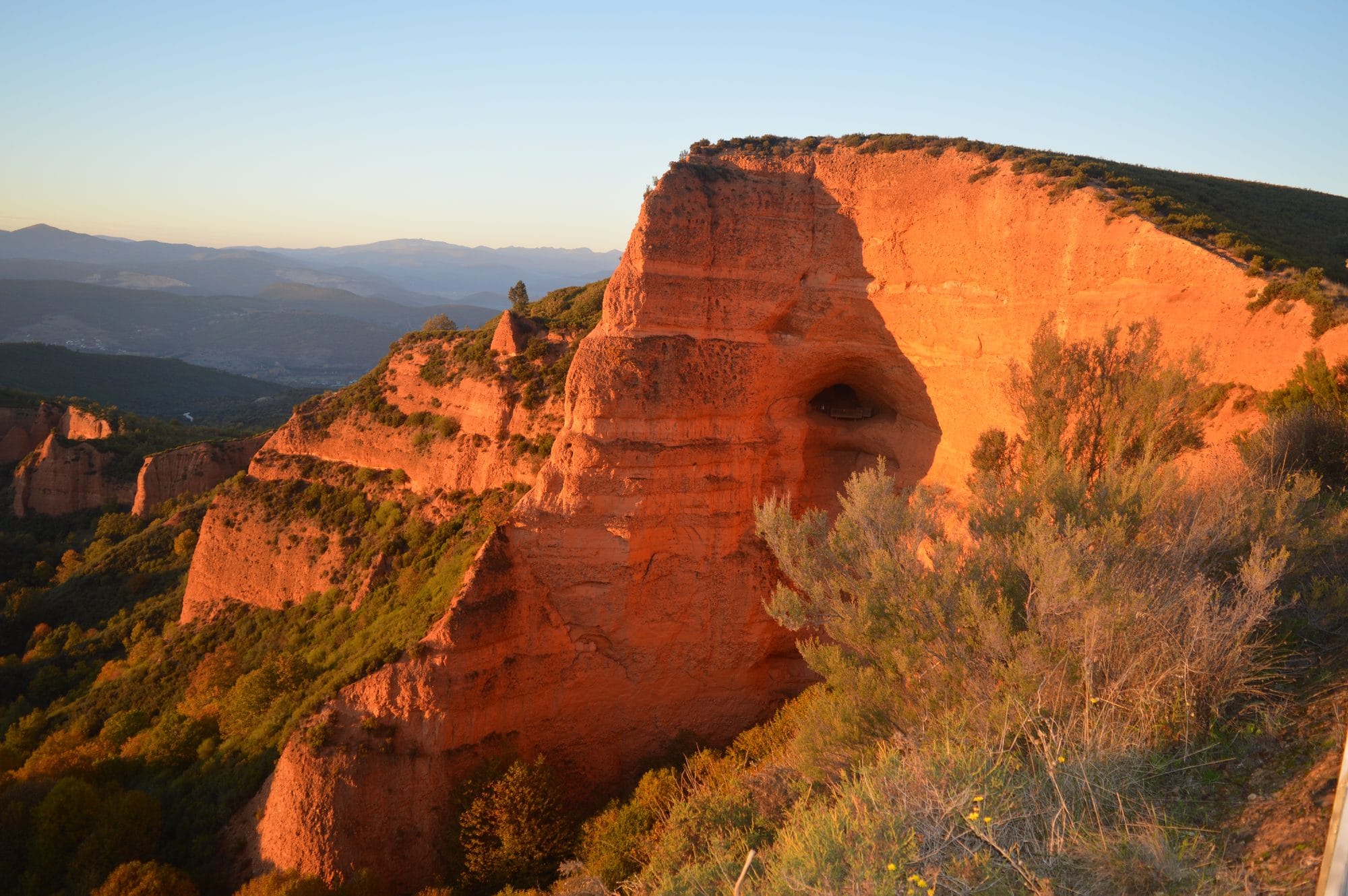 Red mining faces and a dark opening at Las Médulas above wooded terrain in evening sunlight.