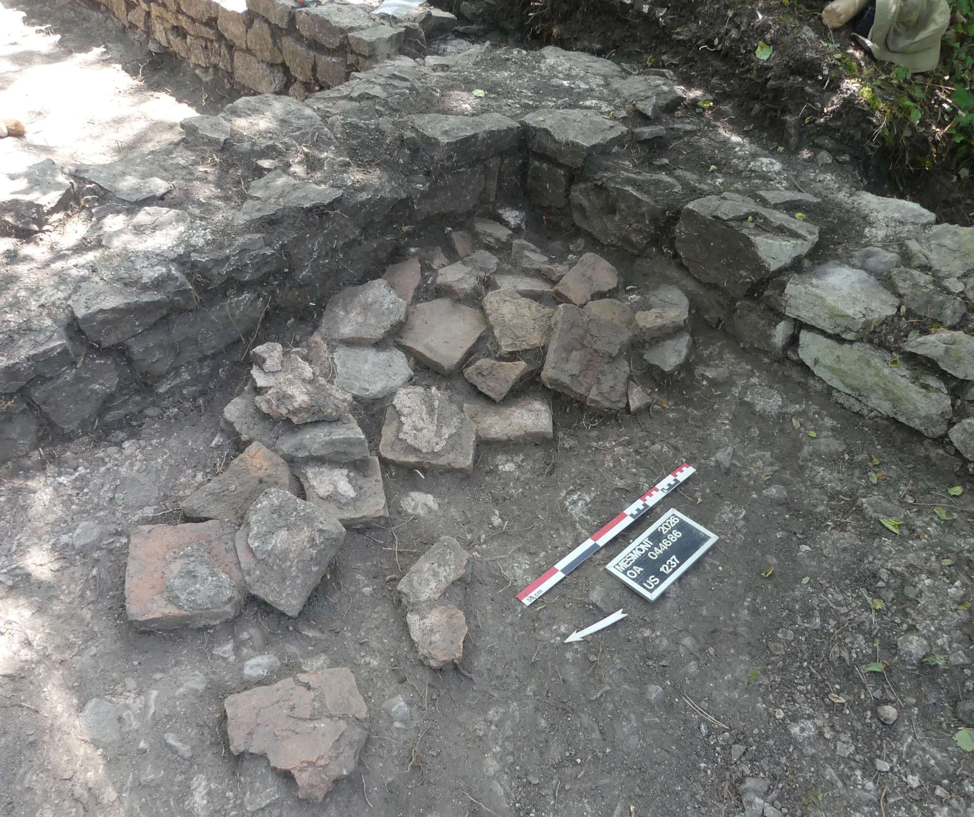 Hypocaust pillar pieces and broken suspended-floor material beside a stone wall at Mesmont.