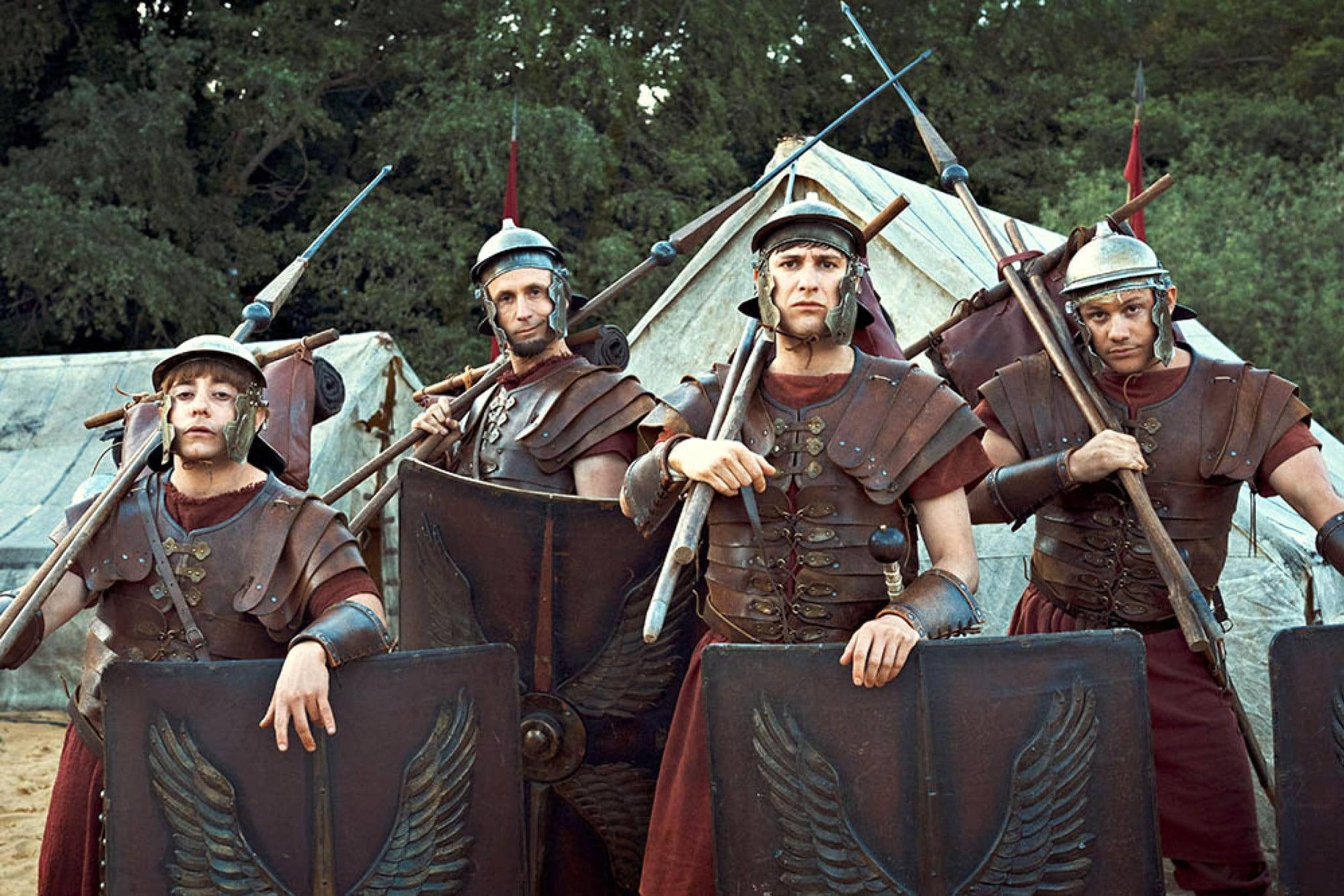 Four Plebs cast members in Roman military costumes, holding shields and spears.