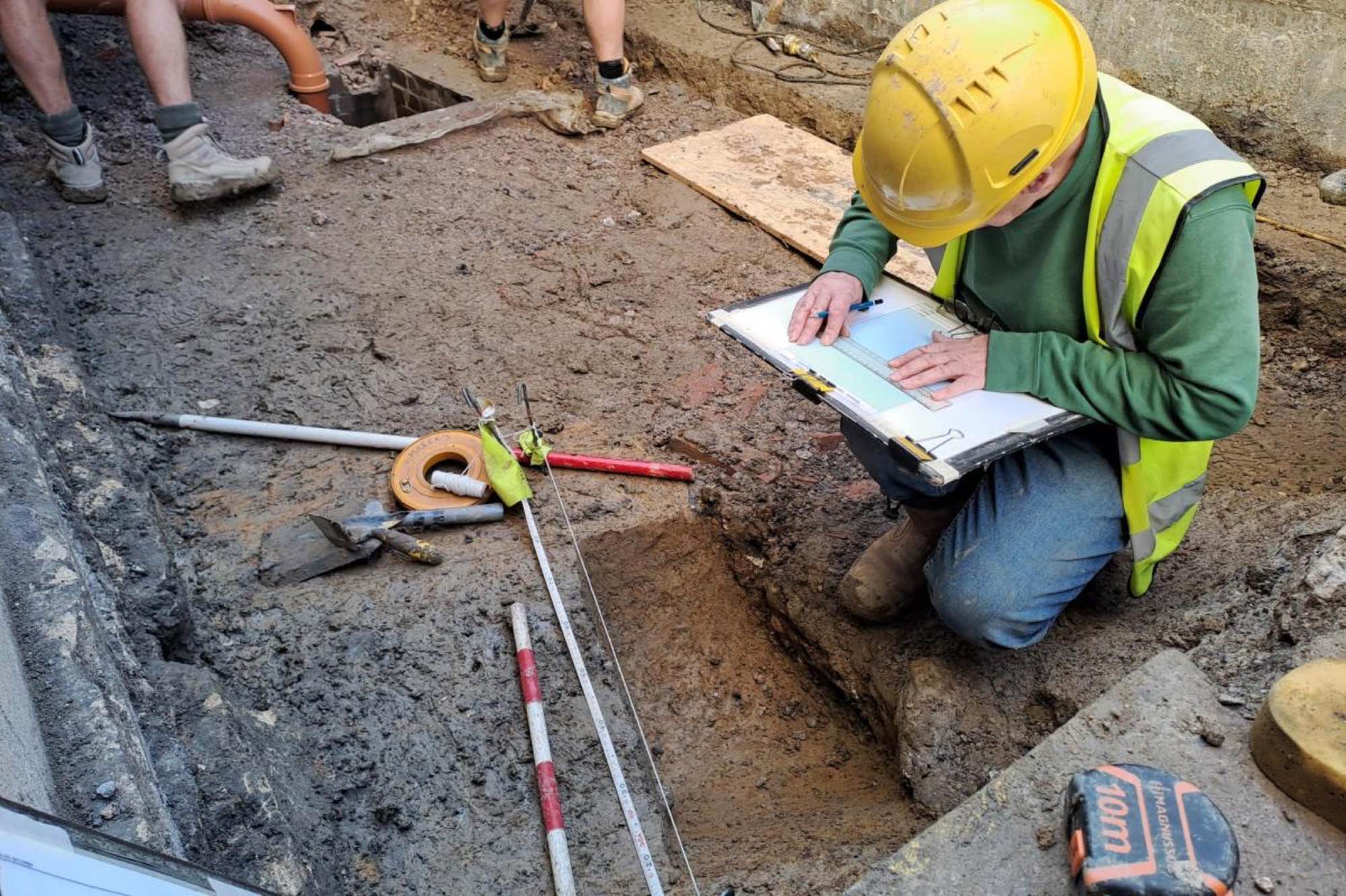 An archaeologist in a yellow hard hat records a Roman ditch in the excavated ground beneath Bath Assembly Rooms.