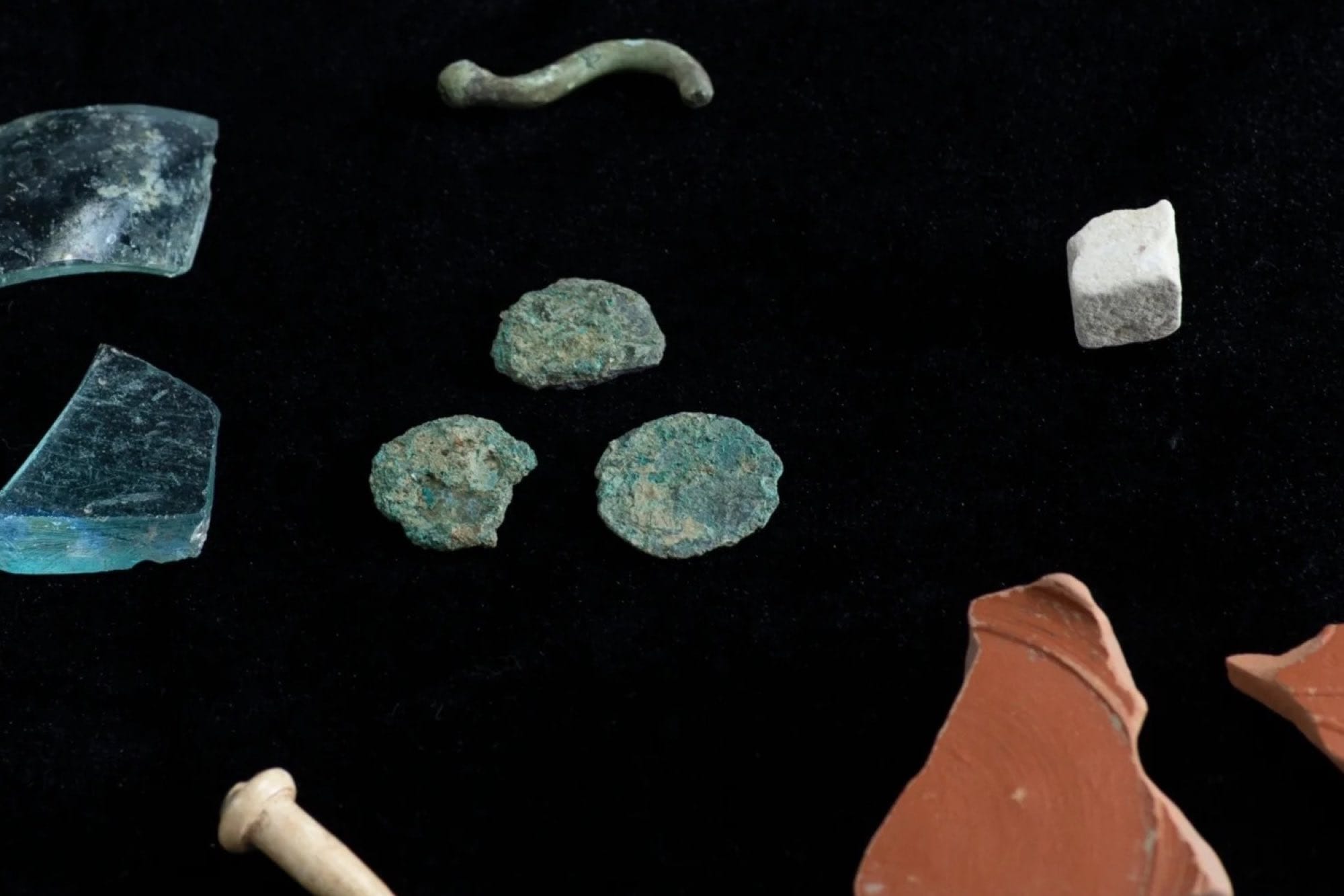 Close view of glass and reddish pottery fragments, rounded corroded objects and a pale stone piece on a black background.