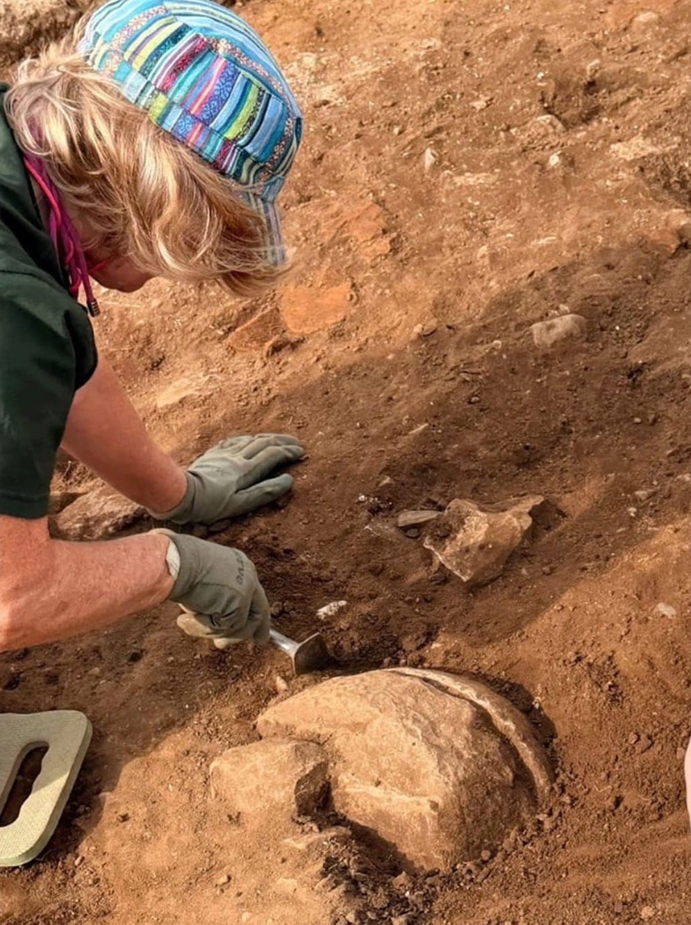 Archaeologist uncovering a circular quern stone in a soil layer at Rossett Roman villa.