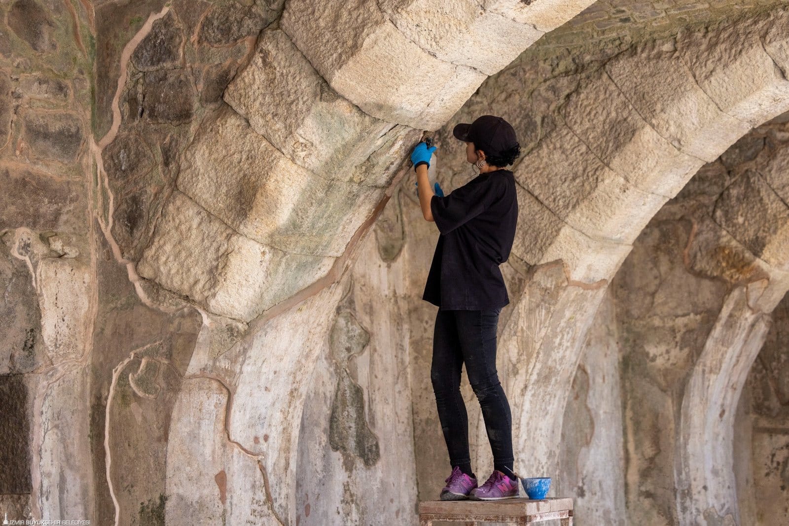 A conservator wearing blue gloves treats surviving plaster beneath a stone arch at Smyrna’s agora.