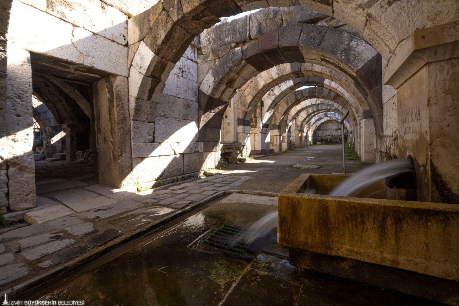 Flowing water in a shallow stone channel beneath successive arches at Smyrna’s agora.