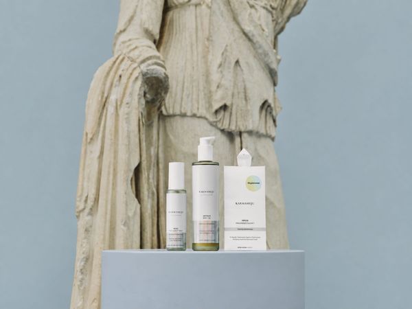 Karmameju ROSE mist, ANTIQUE body oil and VIRIDIS sachet on a pale plinth before an ancient statue.