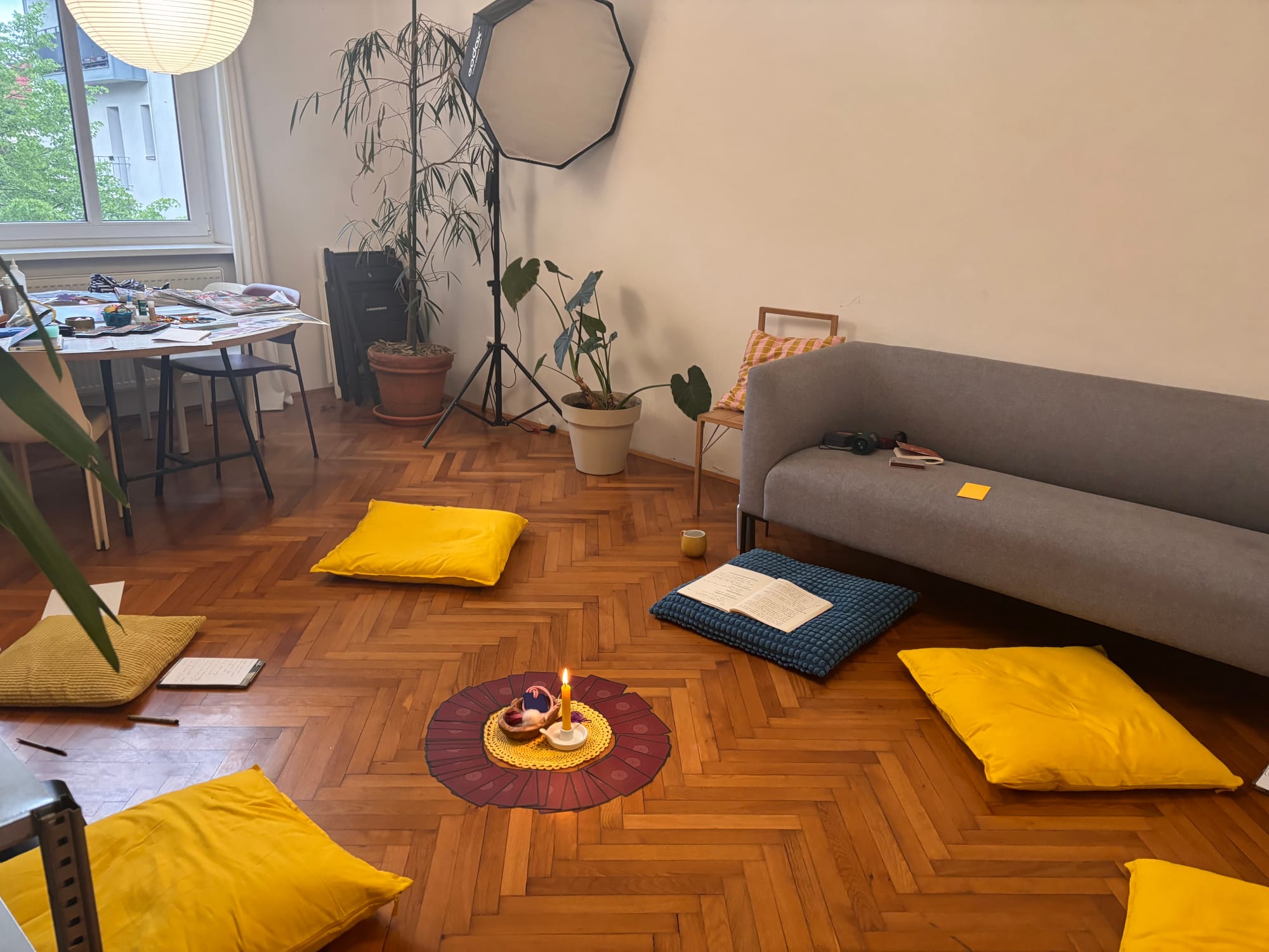 A picture of an office space with pillows on the floor arranged in a circle. in the middle there's a lighted candle with oracle cards spread around it. In the background there is a grey couch, and a big table with some art supplies.