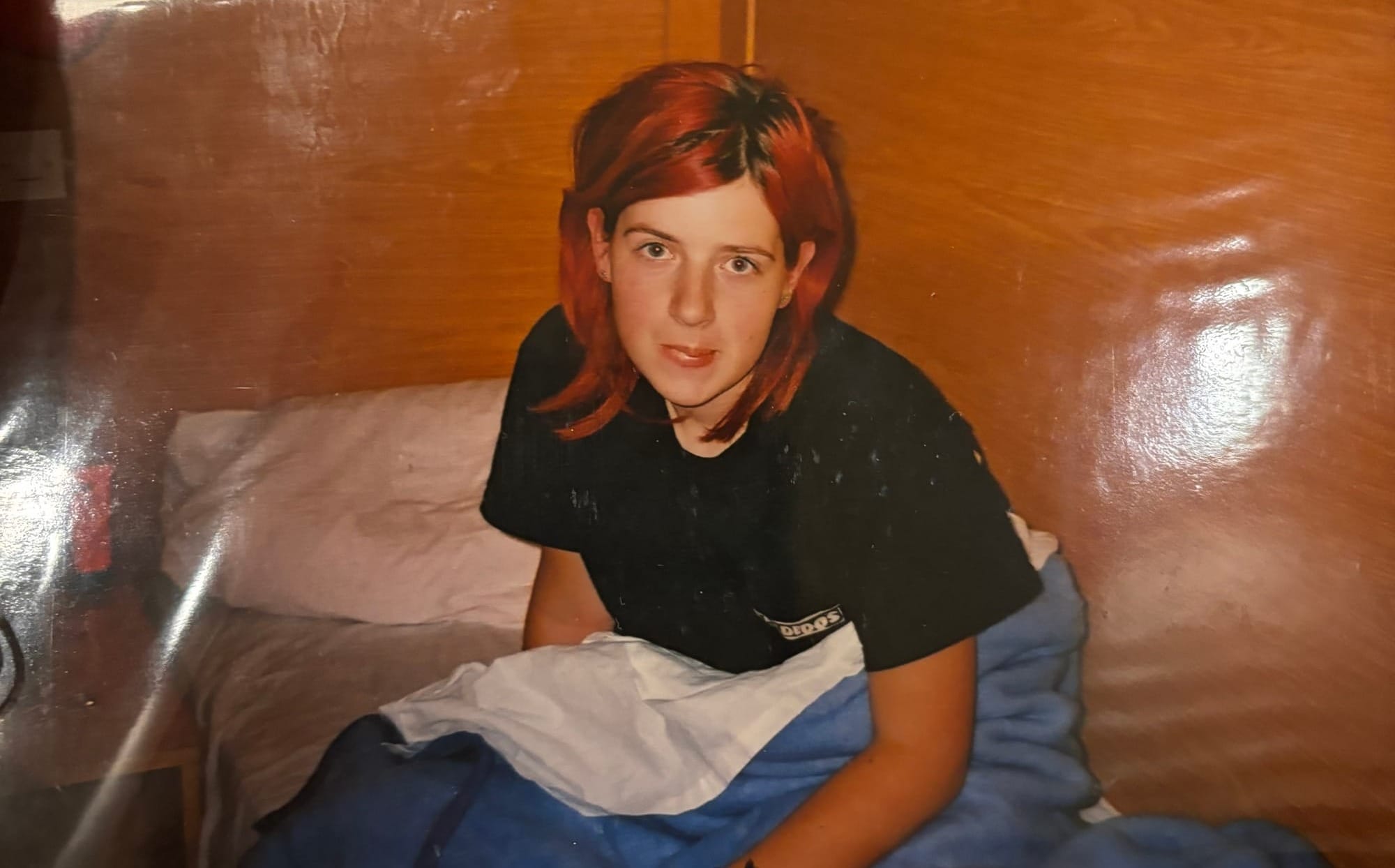 This is a photo of an old picture of the author in high school. A girl with red hair, sitting up in a bed on a school trip. Behind and on the right is a wooden wall, she's wearing a black t-shirt and is covered with a blue blanket.