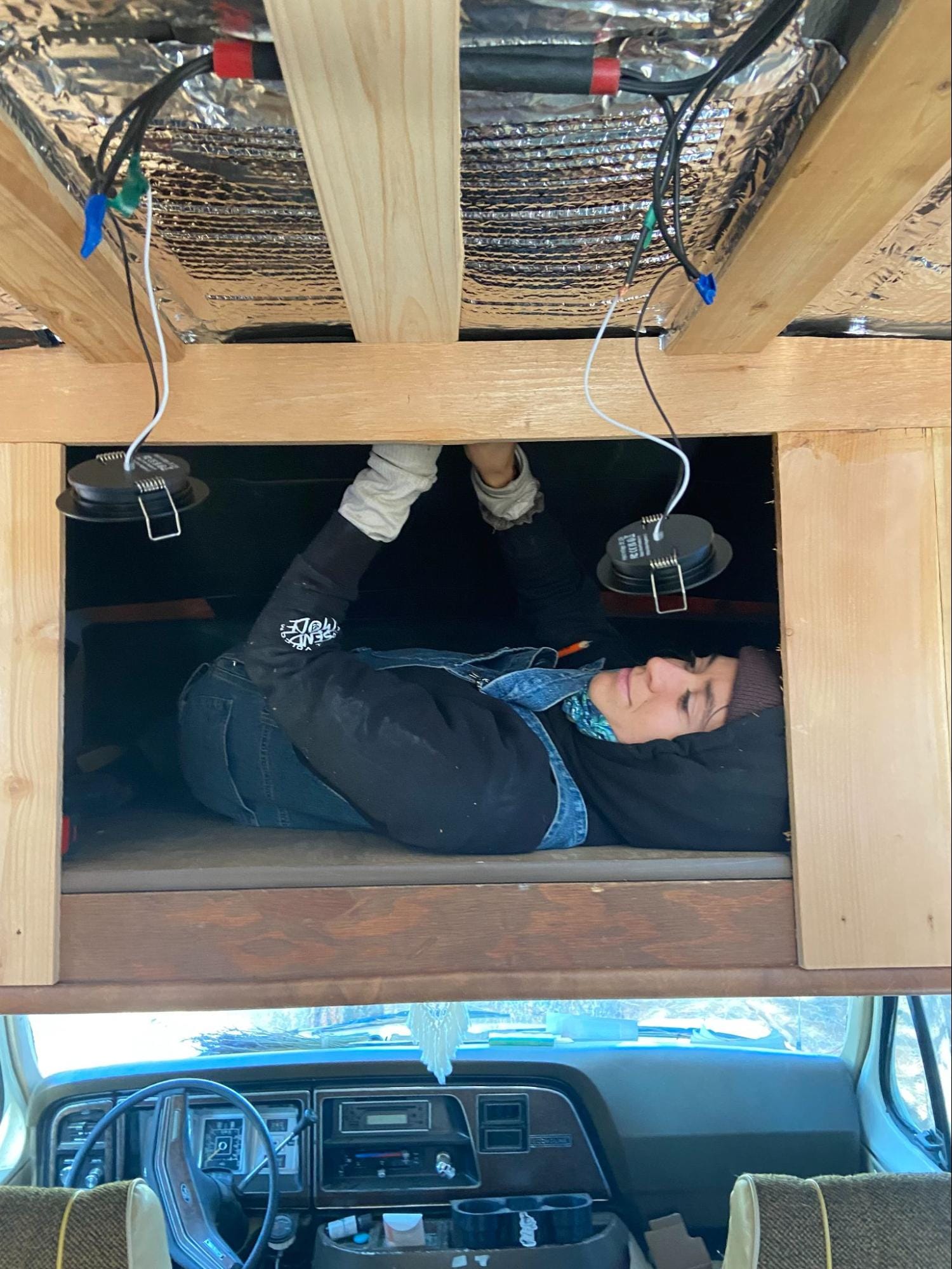 Warmly dressed woman lying on her back installing electrical equipment into a campervan
