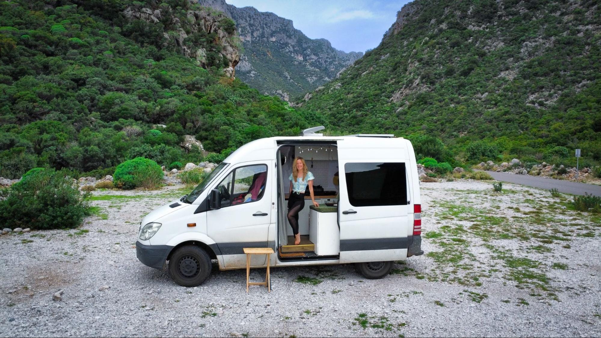 Young woman standing in the side doorway of a campervan parked in a canyon surrounded by lush rocky cliffs