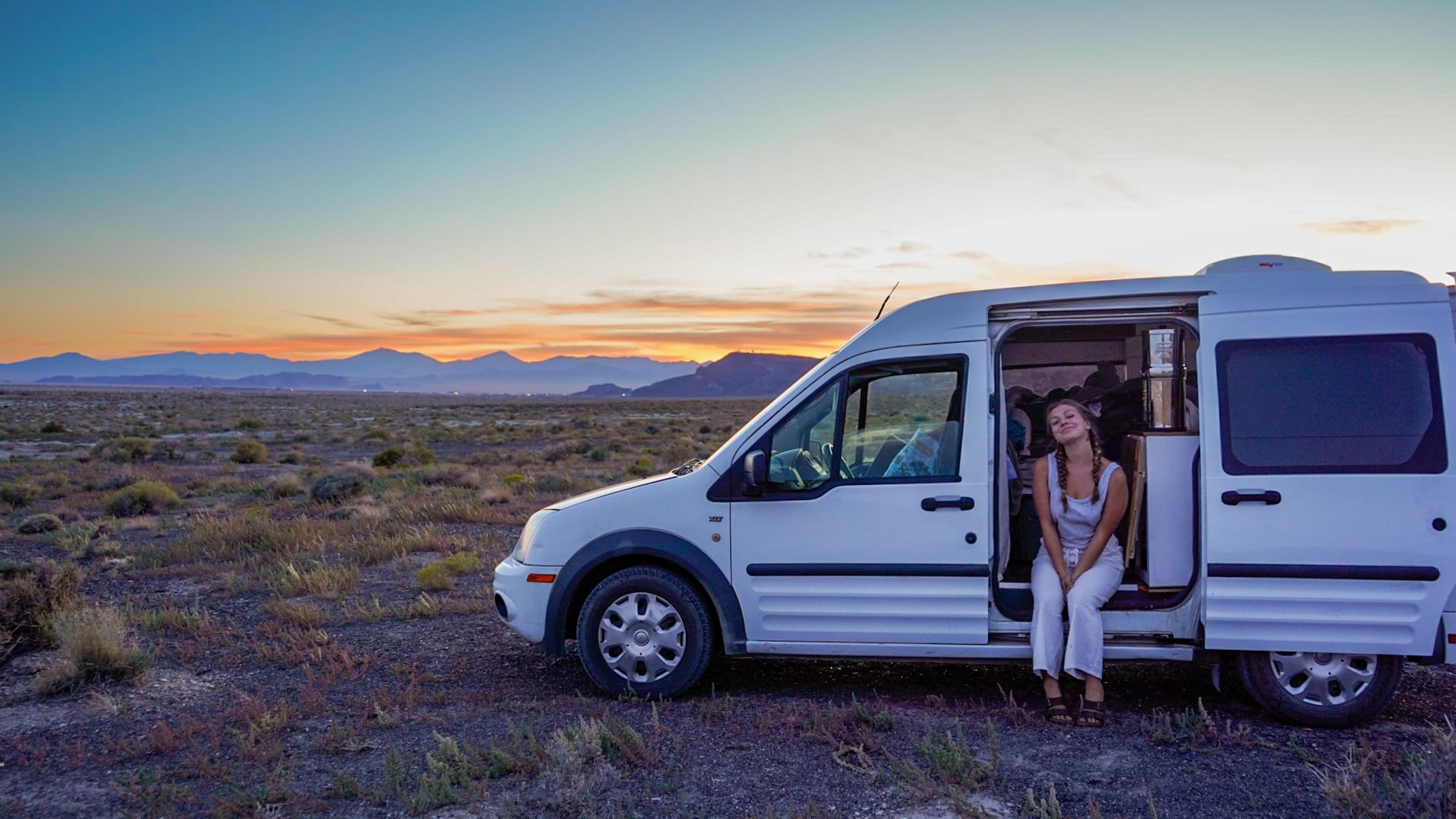 Young woman sitting in the side doorway of a compact campervan in a semi desert wilderness at dusk
