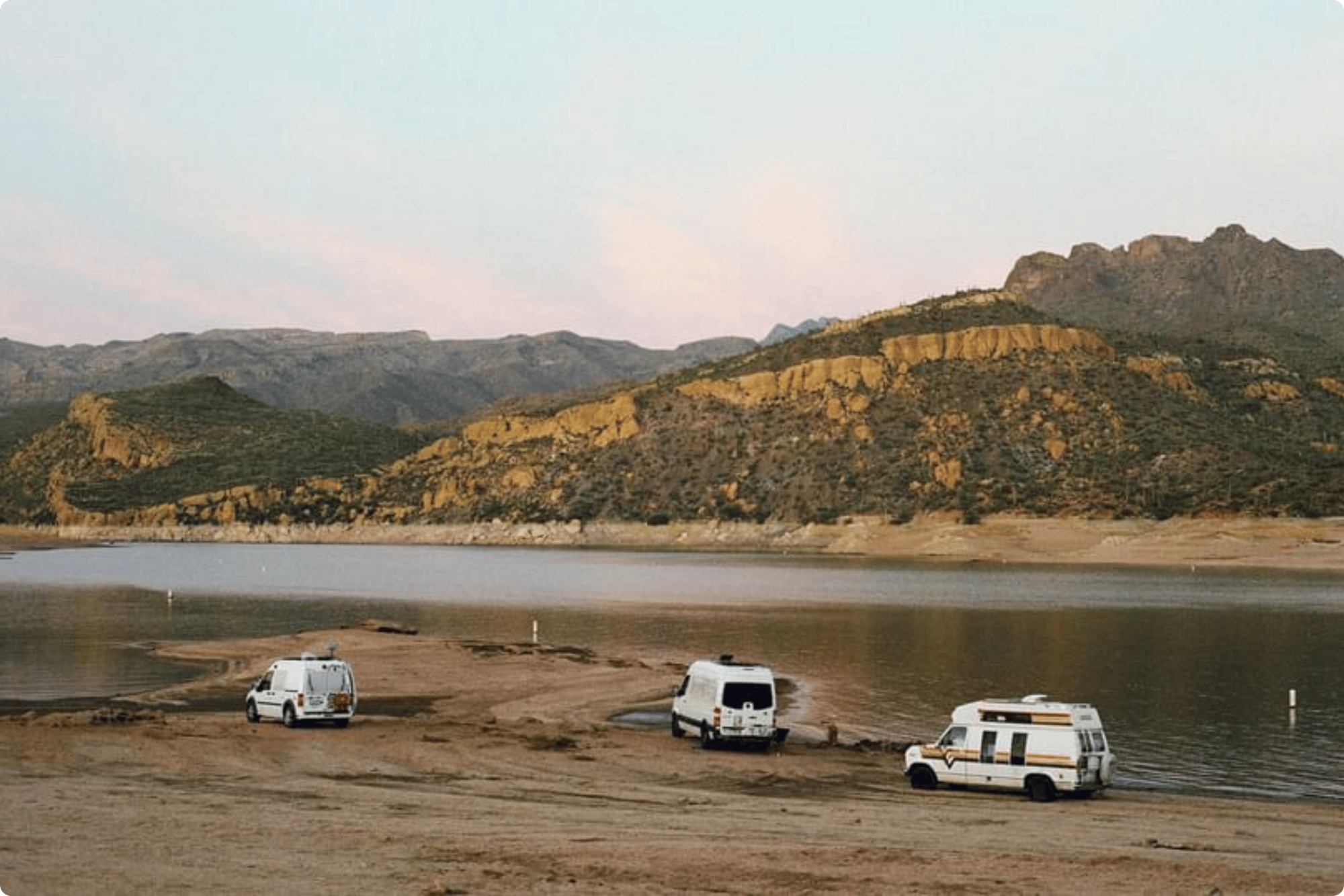 Three campervans parked near a lake with mountains in the distance