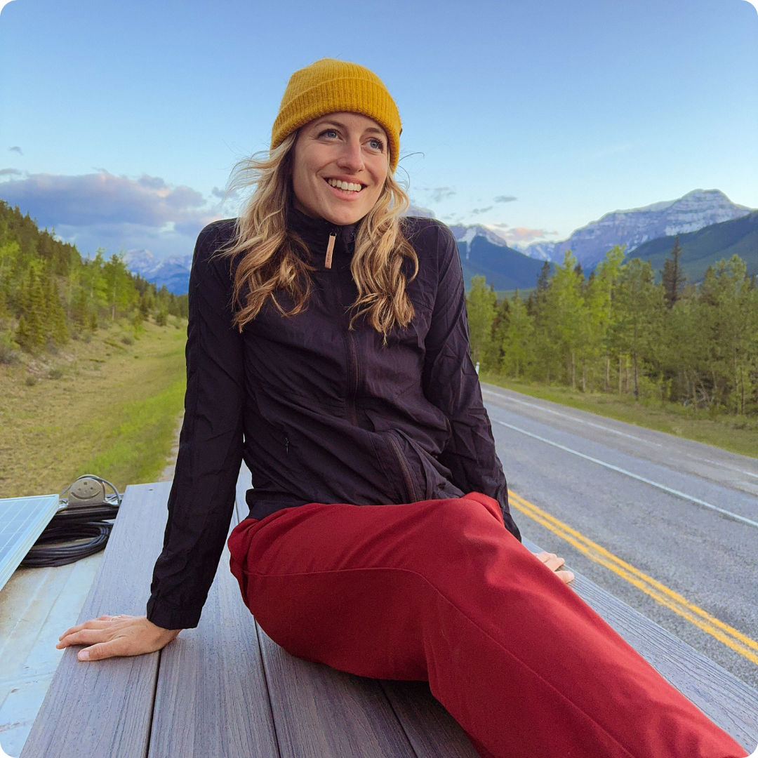 Girl sitting on the roof of a campervan with the road and mountains in the background
