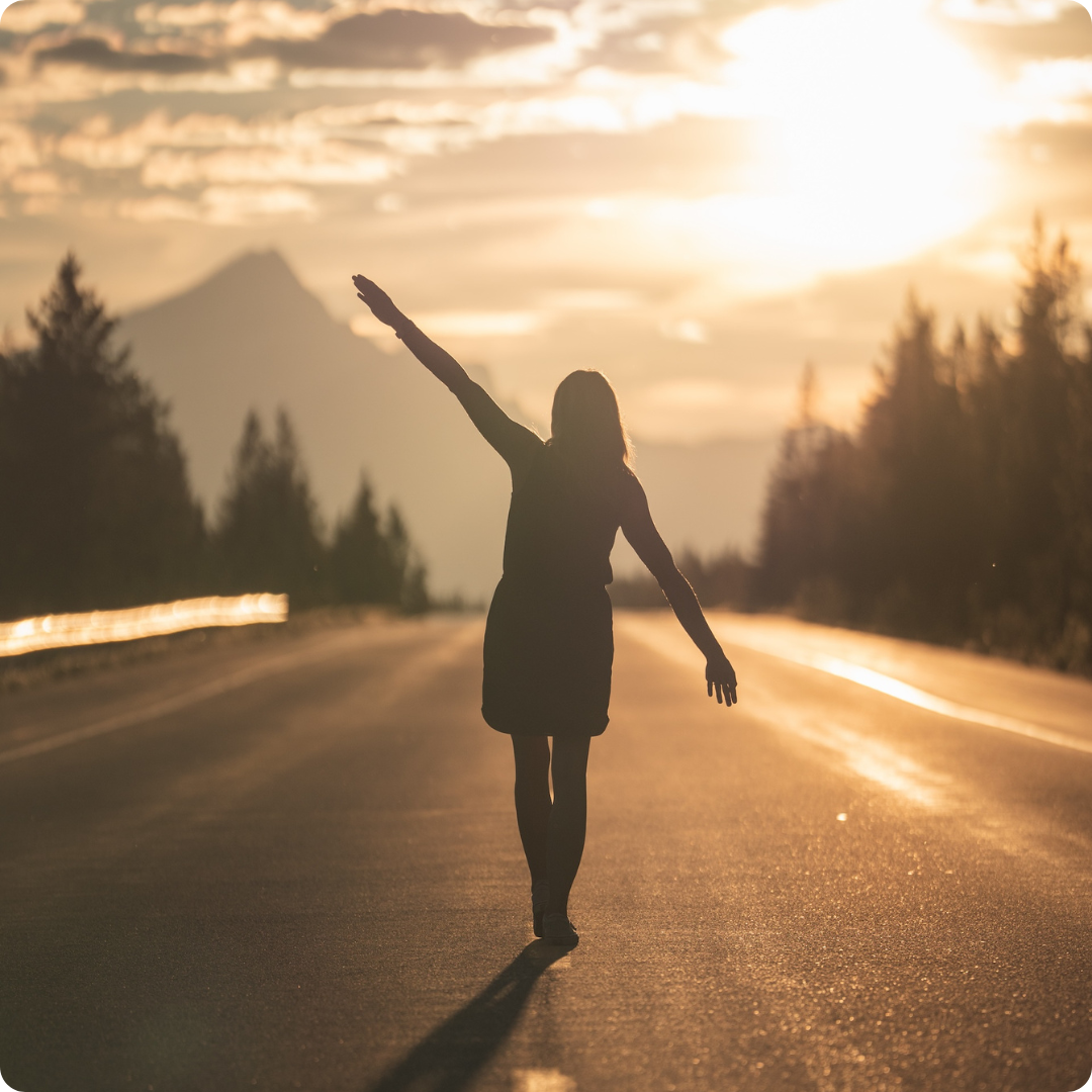 Girl walking down the middle of an empty road in the wilderness at dusk with sunsetting over mountains in the distance