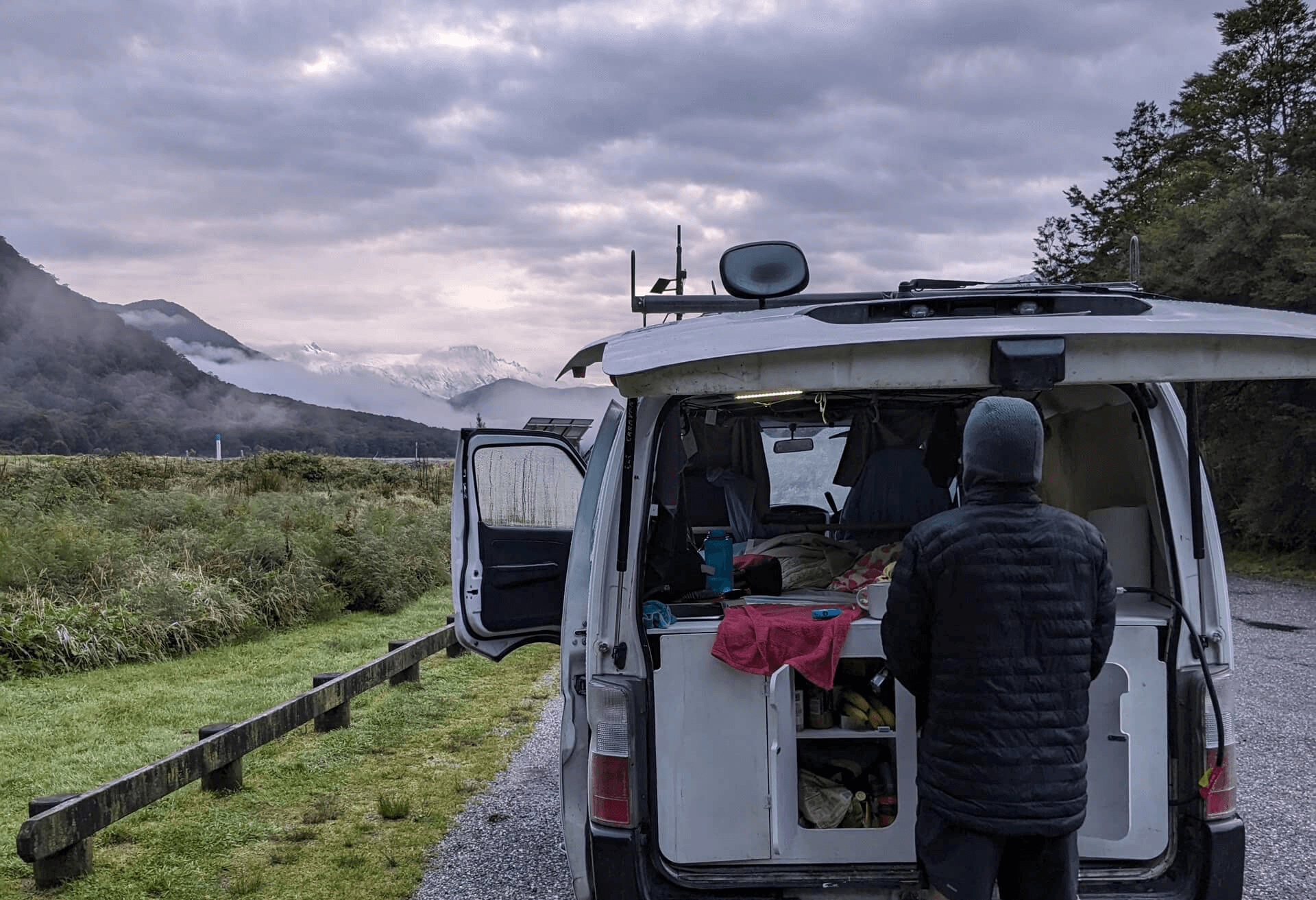 Camervan parked on the side of a road with snow capped mountains in the distance and a man preparing food at the back of the van