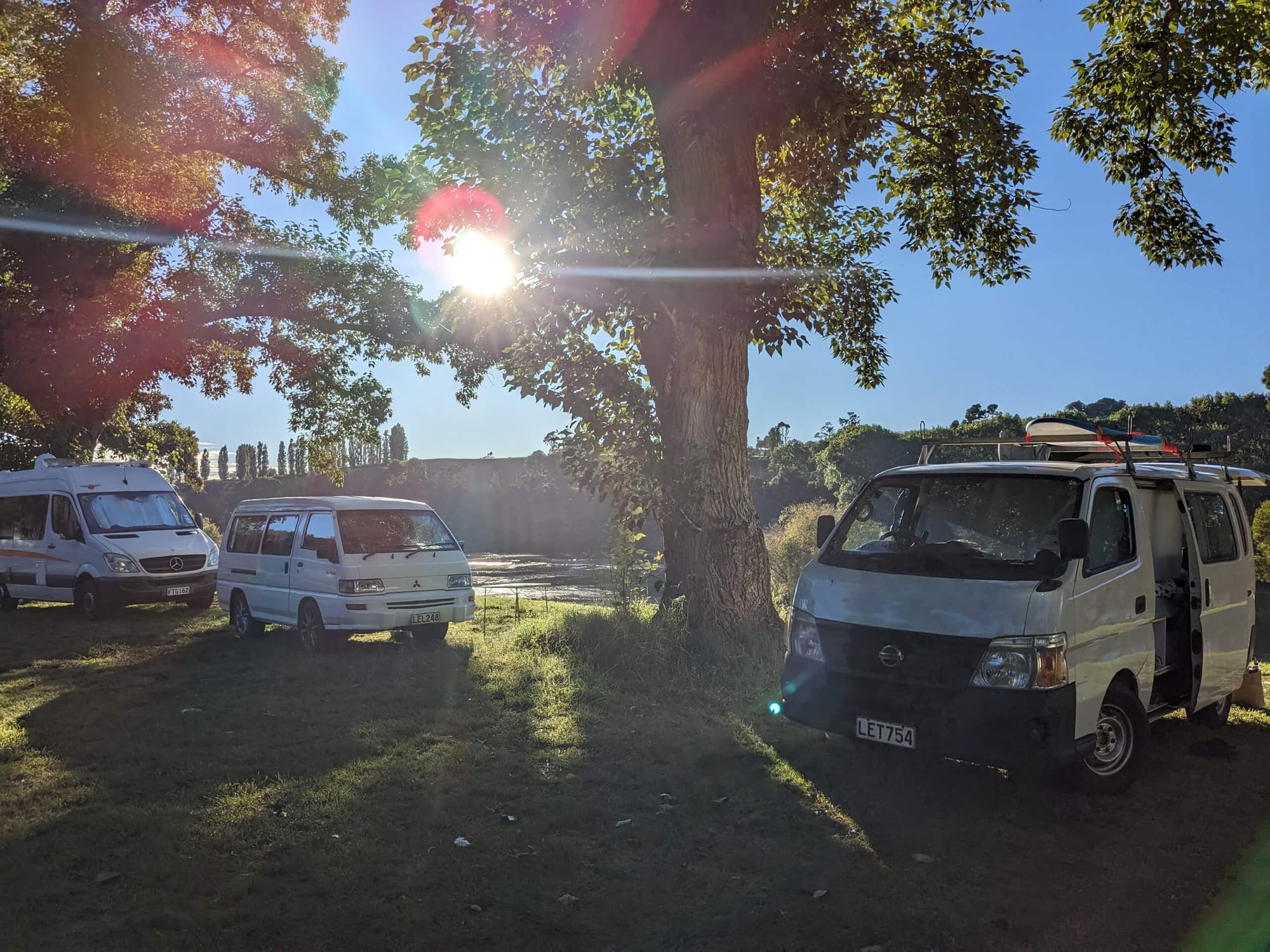 Three white campervans parked under a tree next to a river with the sun shining through the trees