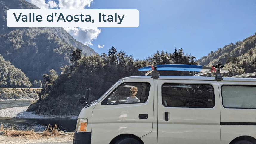 White campervan with blue surfboard on the roof in a mountainous region of Italy