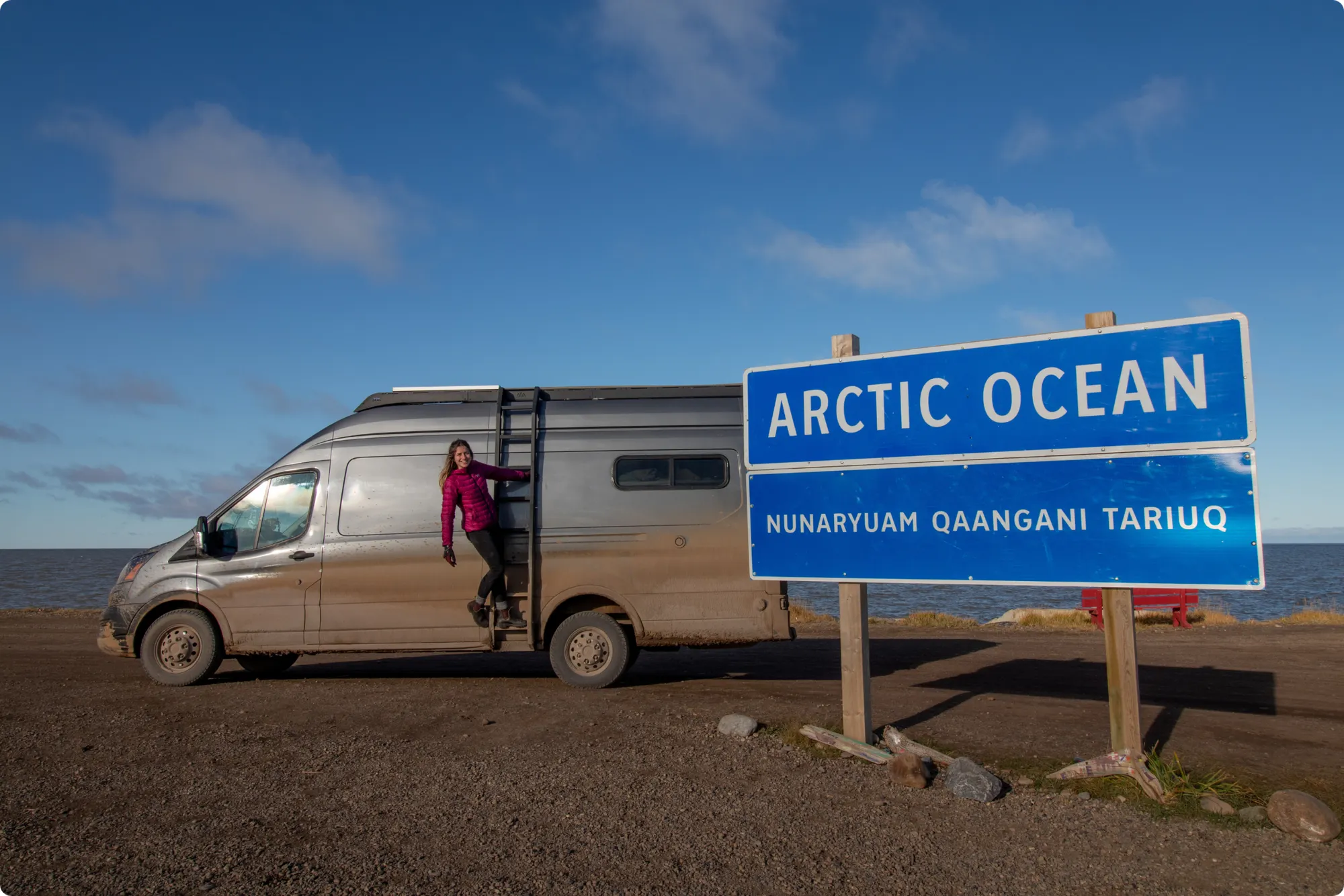 Woman standing on campervan side ladder next to a sign that reads "Arctic Ocean, nunaryuam qaangani tariuq"