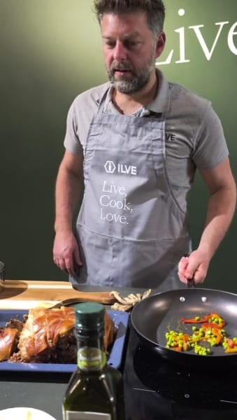 Man cooking peas in a pan with sizzling pork on counter.