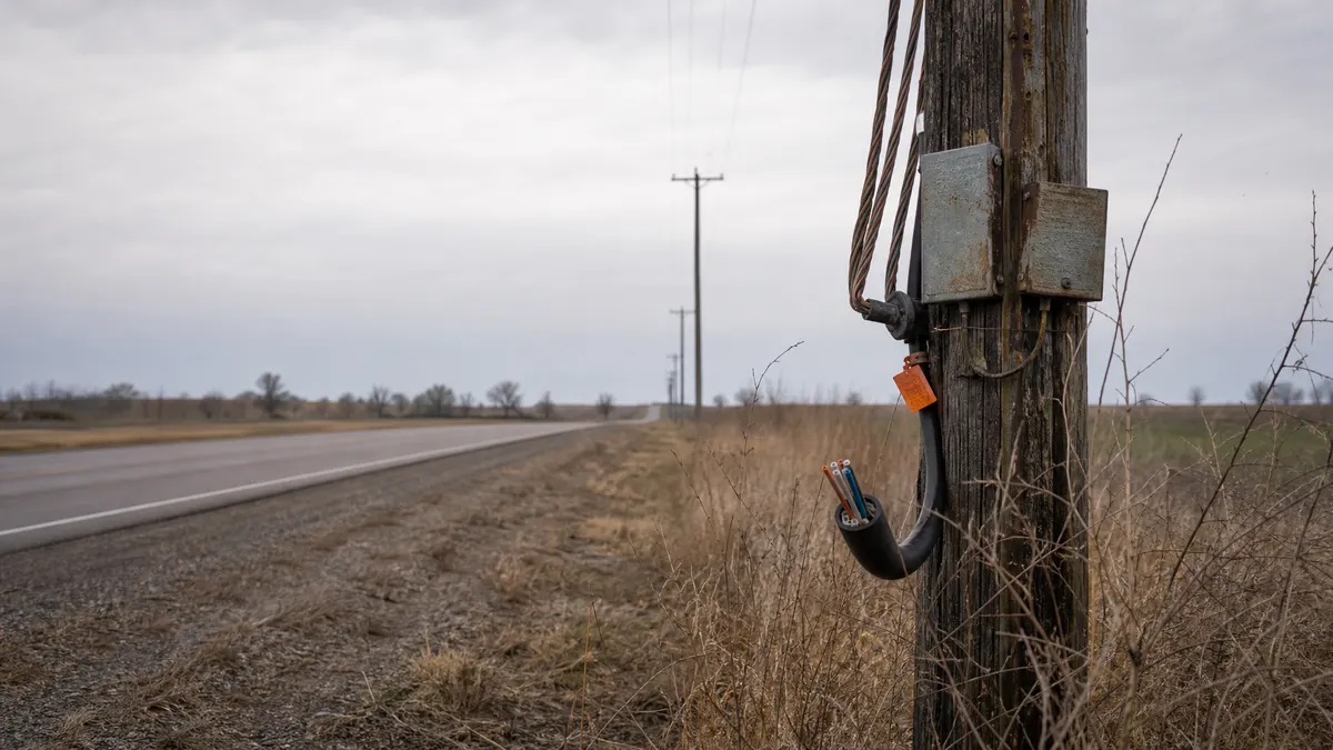 A rural telephone pole with old copper lines, repair tags, and a fiber conduit stopping short nearby, showing legacy network costs during a technology transition.