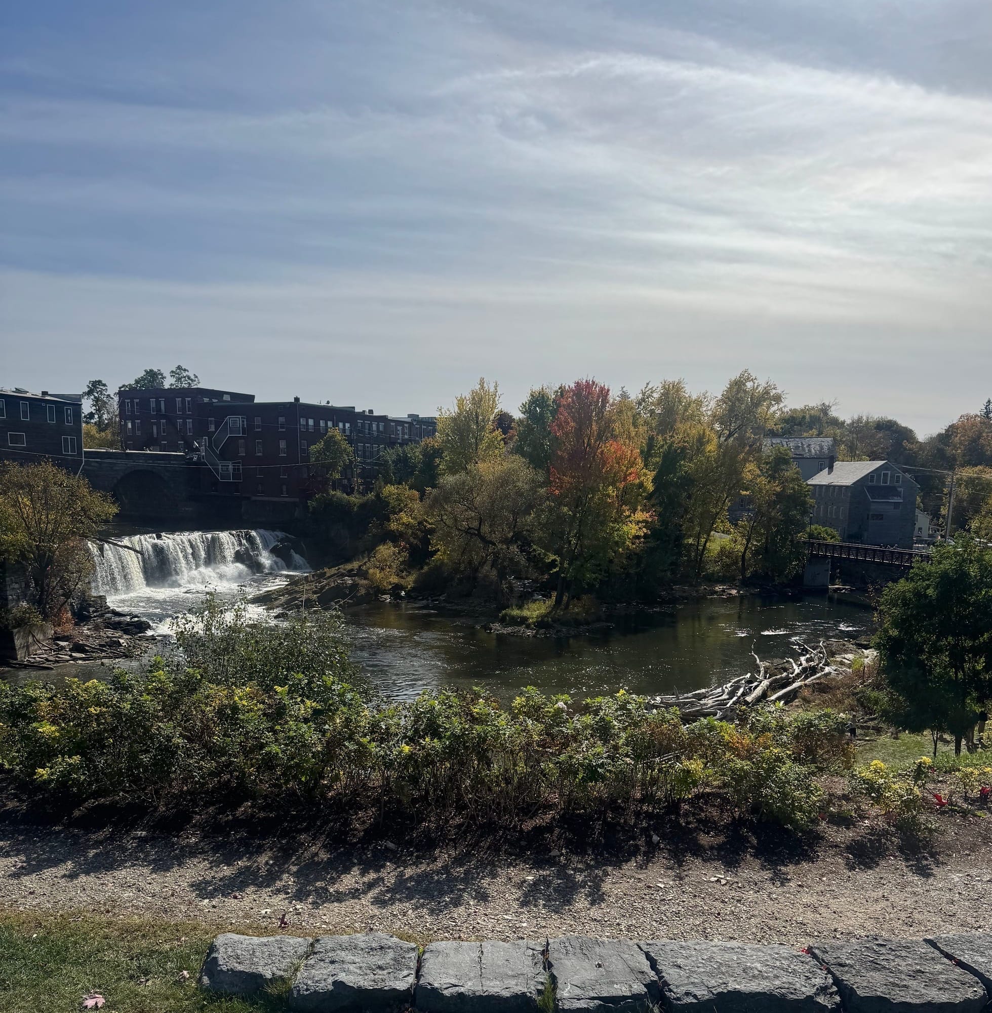 A waterfall flows through the heart of Middlebury, Vermont, surrounded by historic buildings and trees beginning to show their peak autumn colors under a cloudy blue sky.