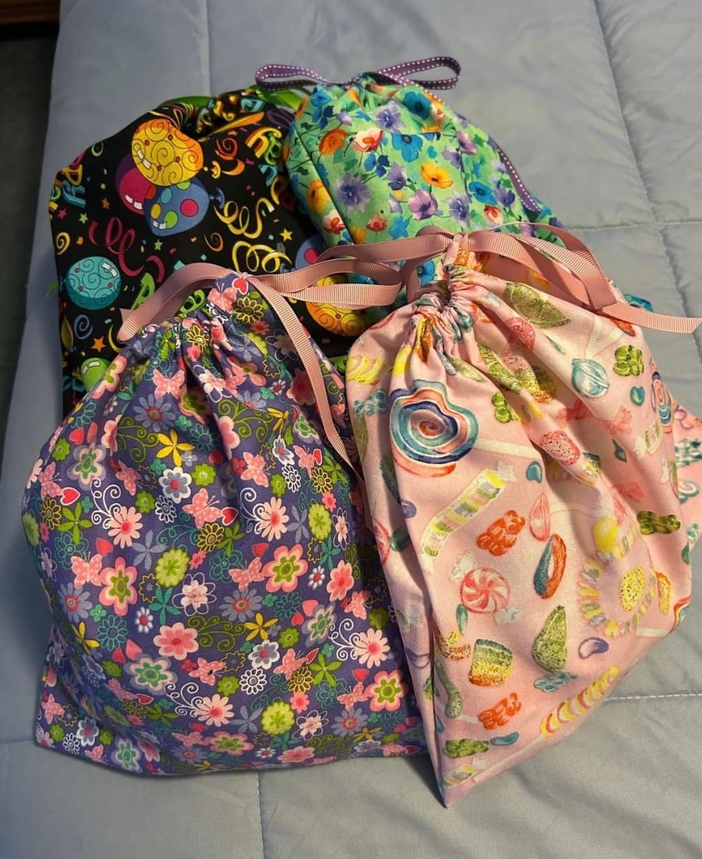 Four colorful handmade fabric drawstring bags with floral, candy, and whimsical patterns, gathered together on a light gray quilt.
