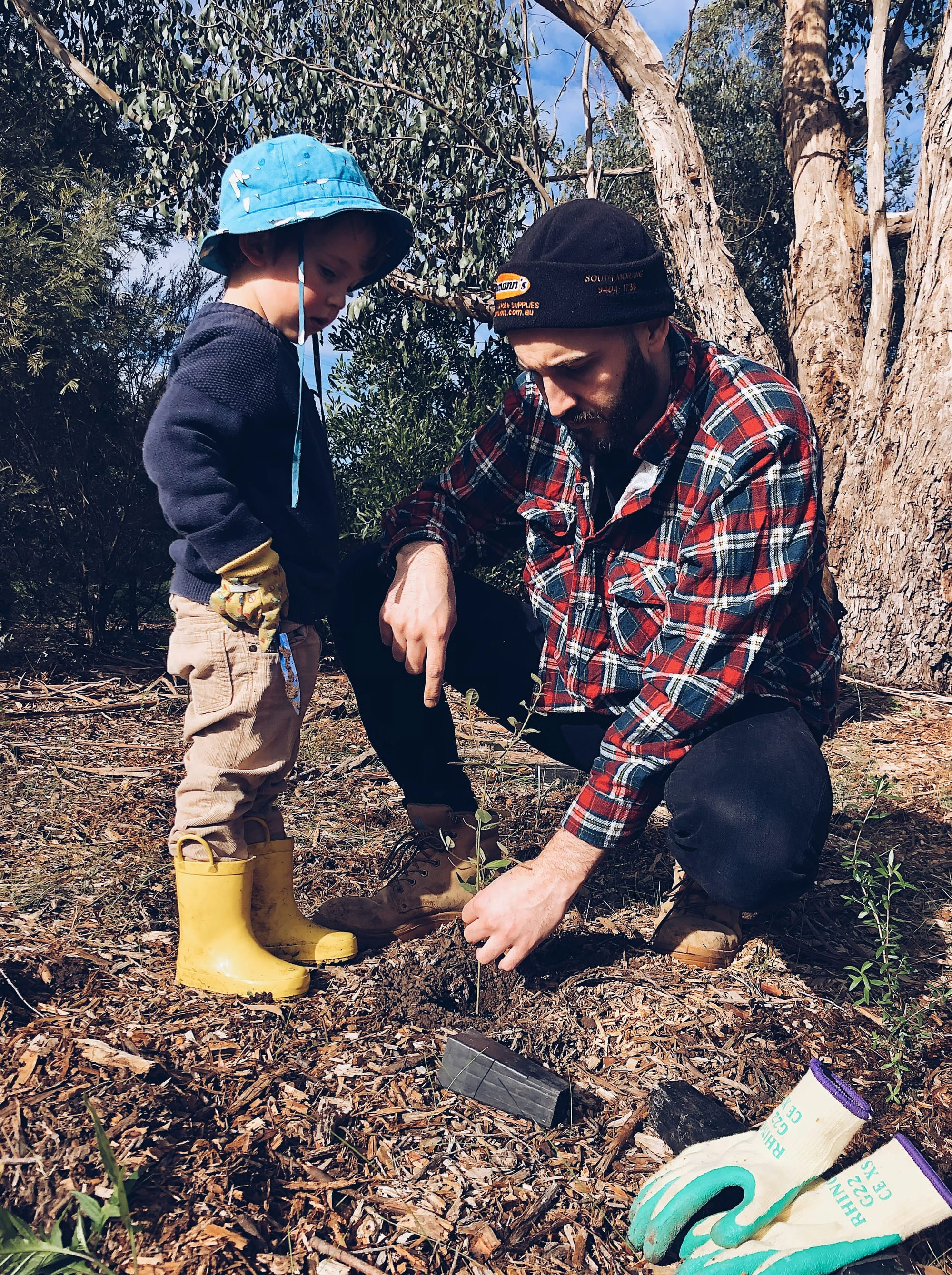 A father kneels beside his young son as they plant seeds together in the soil.
