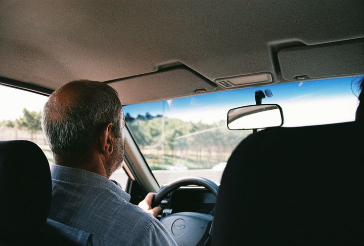 A man driving a car, seen from the back seat, looking out at the road ahead.