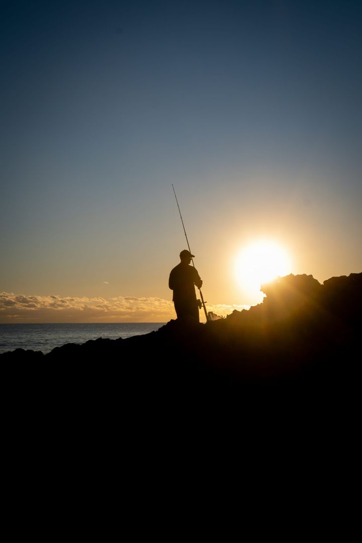 Silhouette of a fisherman standing on rocky shoreline with the sun low over the ocean, holding a fishing rod and looking out at the water.