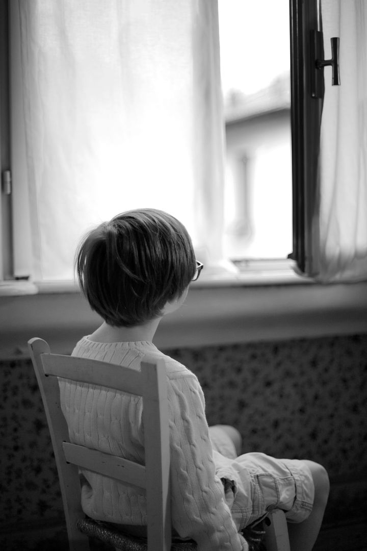 Black-and-white photo of a child sitting quietly by a window, appearing to wait for someone to come home.