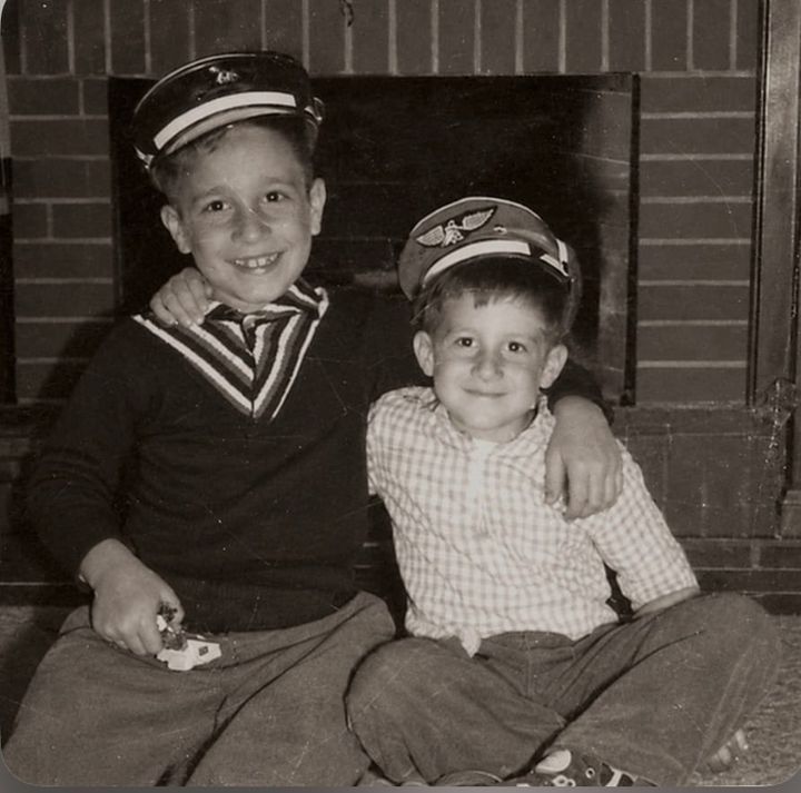 Two young brothers sitting close together in front of a fireplace, wearing Air Force hats, with one brother’s arm around the other.