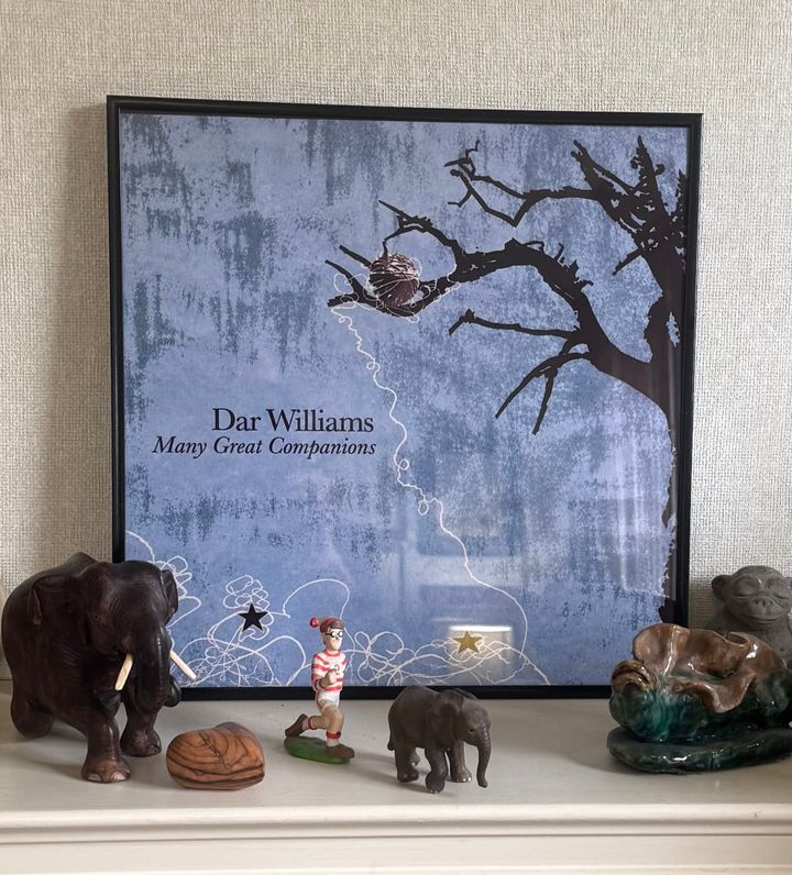 A framed cover of Dar Williams’ Many Great Companions, surrounded by elephant figurines, a small traveler, and other treasured objects on a mantel.