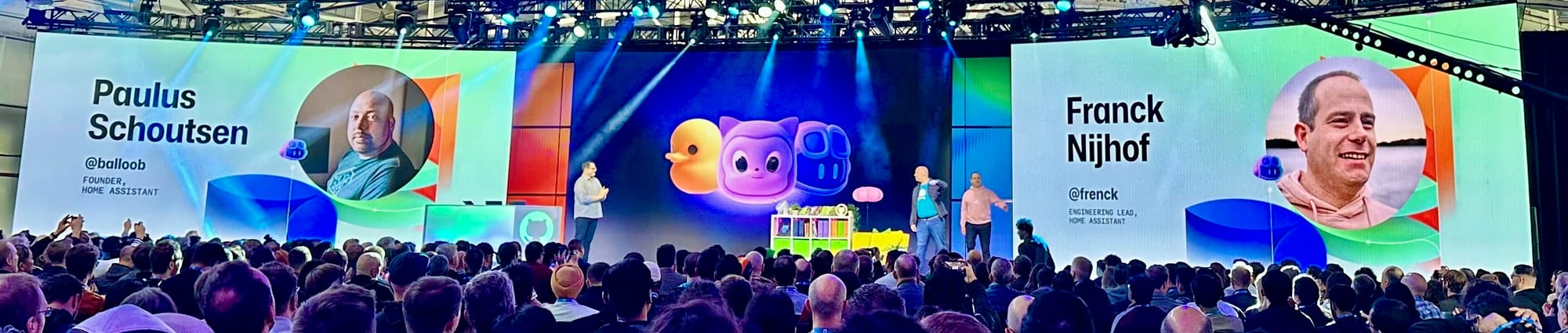 Tech conference crowd watches two speakers onstage. Screens show portraits labeled Founder and Engineering Lead for Home Assistant beside GitHub mascots.