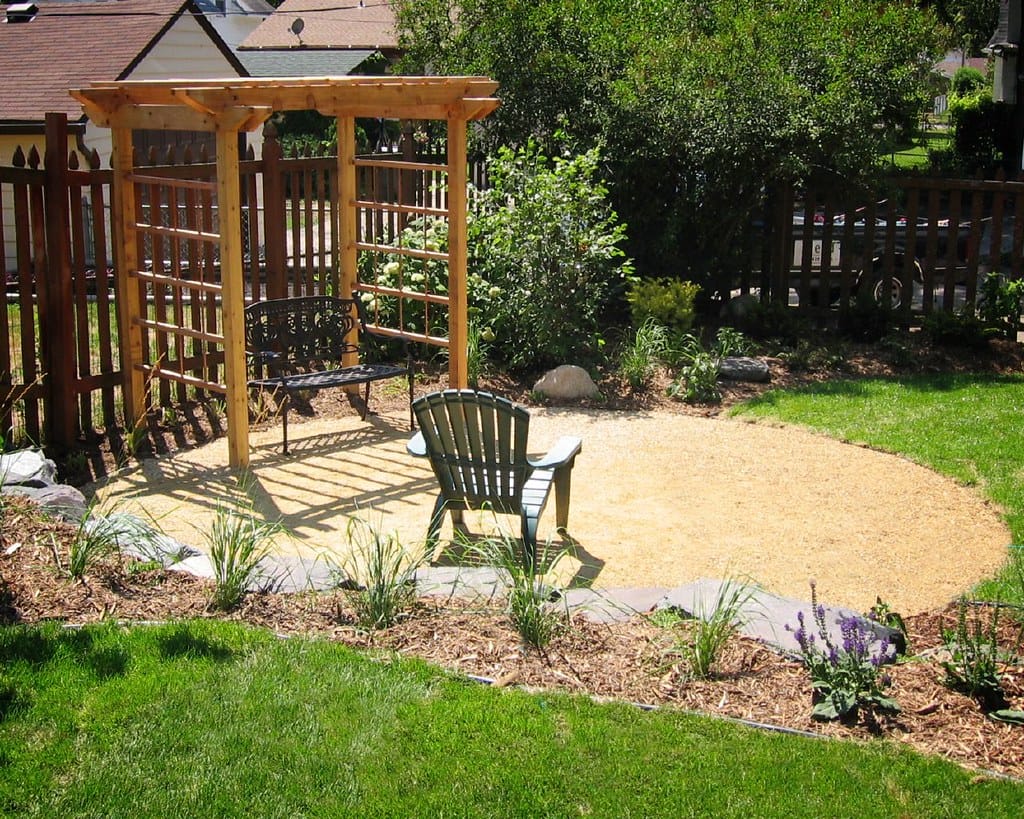 Circular crushed gravel patio with pergola and chairs edged by mulched planting beds