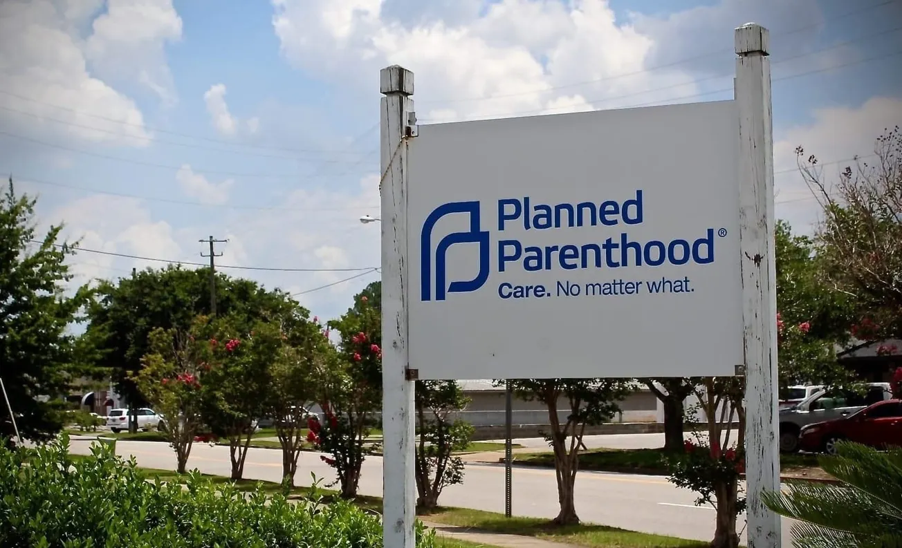 A photograph of a white sign with blue text featuring the Planned Parenthood logo and the slogan "Care. No Matter What." There are green grass and trees and a blue sky in the background.