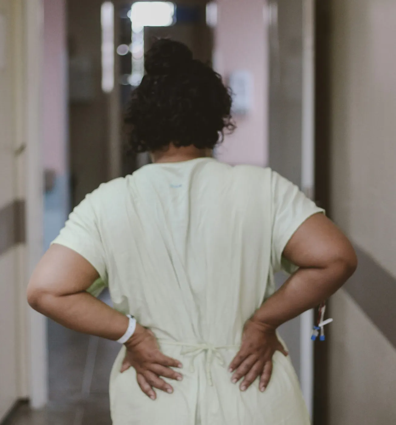 A photograph of a Black woman wearing a white medical gown. Her hands are on her lower back and she is facing away from the camera
