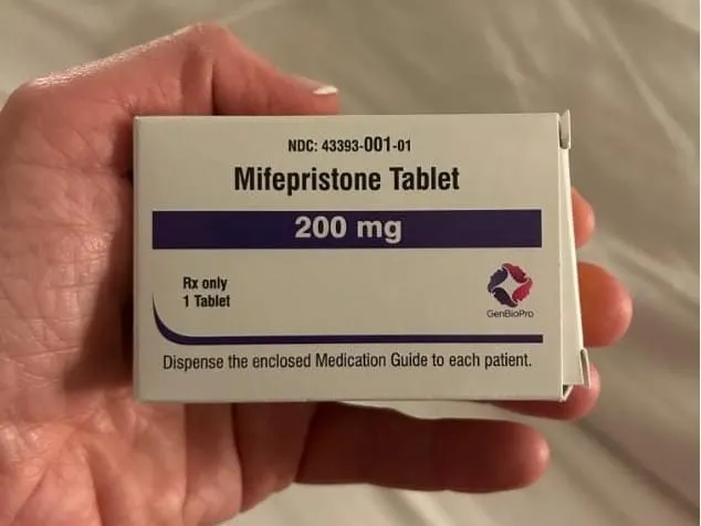A photograph of a white woman's hand holding a mifepristone pill box.